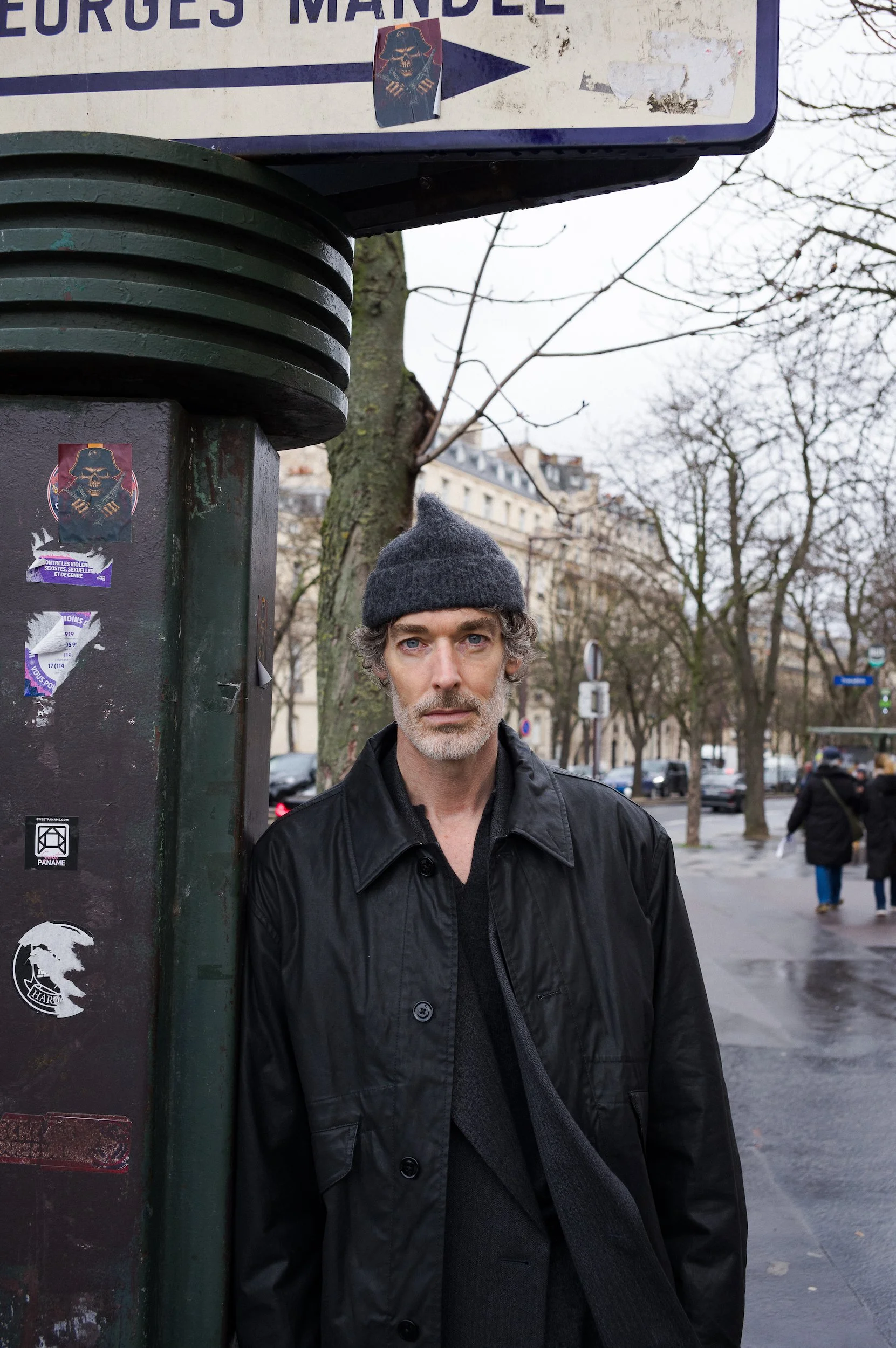 A man with curly gray hair and a beard wearing a black leather jacket and a gray knit beanie standing outdoors on a cloudy day, with leafless trees and buildings in the background. Part of a street sign with stickers is visible on the left side.
