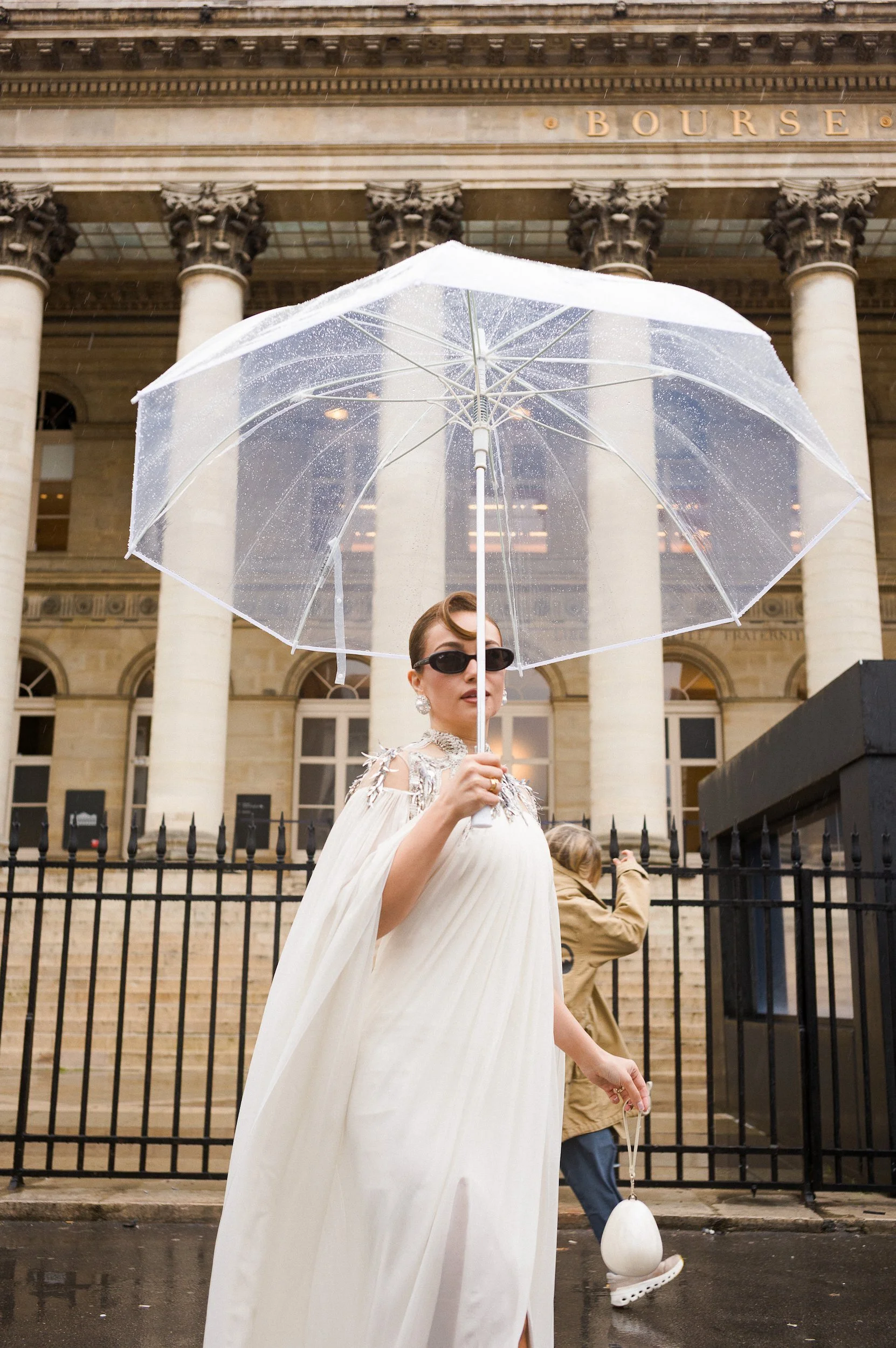 At Paris Fashion Week, Woman in white dress holding a clear umbrella with raindrops in front of the New York Stock Exchange building, with columns and the sign 'BOURSE' visible.