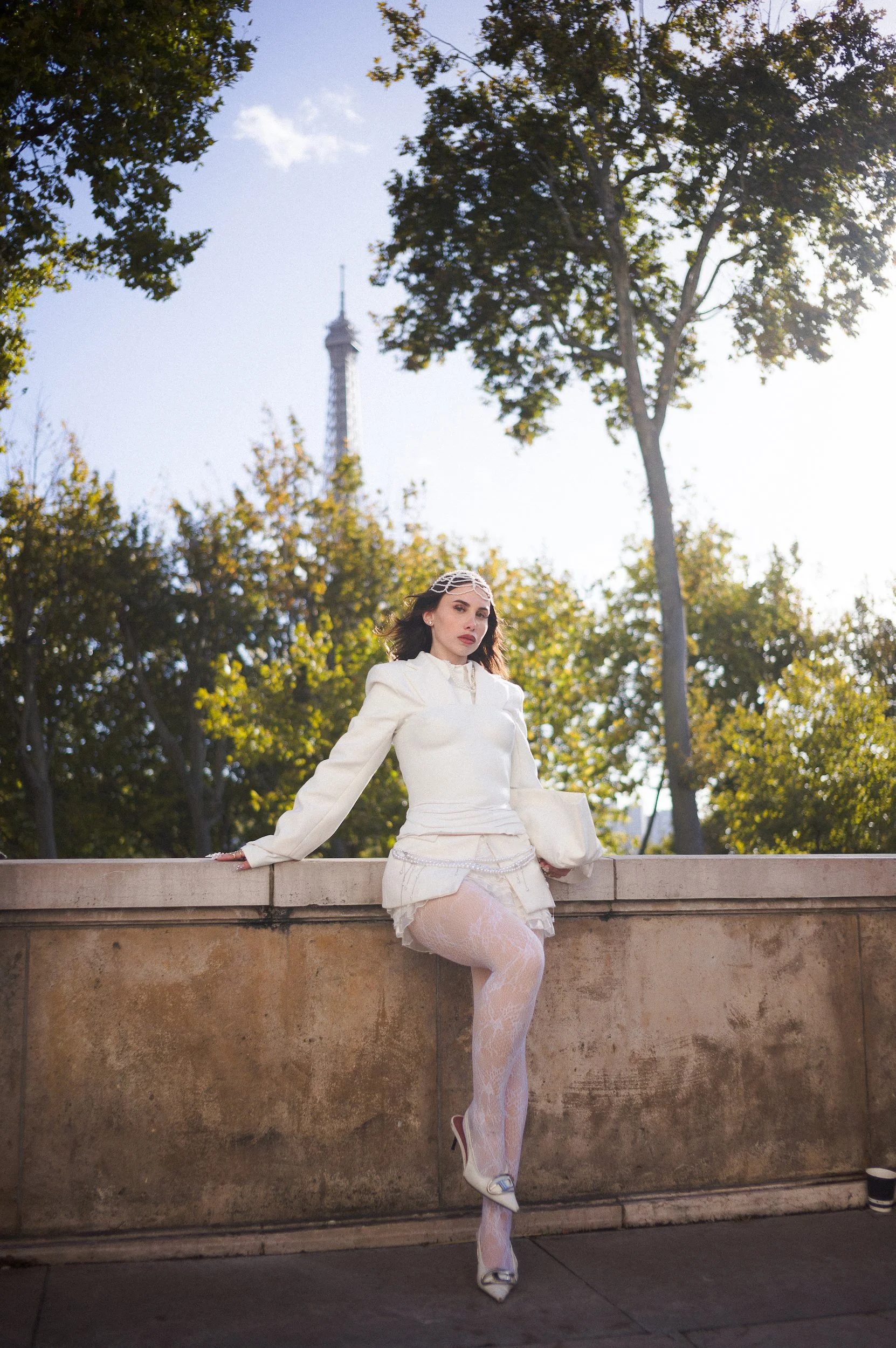 At Paris Fashion Week, Woman in white outfit with lace fashion, sitting on a stone ledge in front of trees, with the Eiffel Tower visible in the background during daylight.