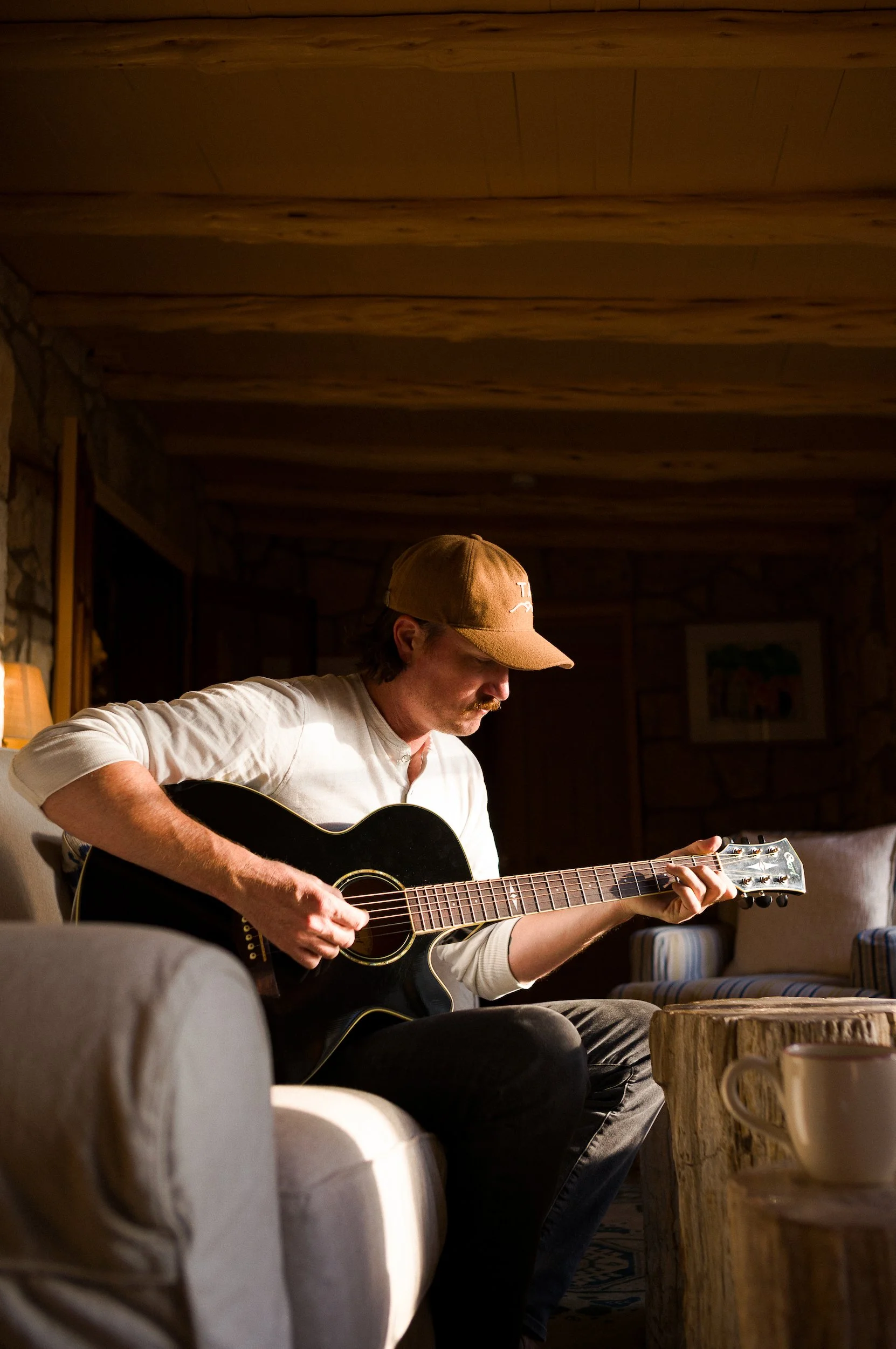 A man playing a guitar in a cozy, rustic room with wooden ceiling beams and stone walls, sitting on a couch with a striped chair nearby.