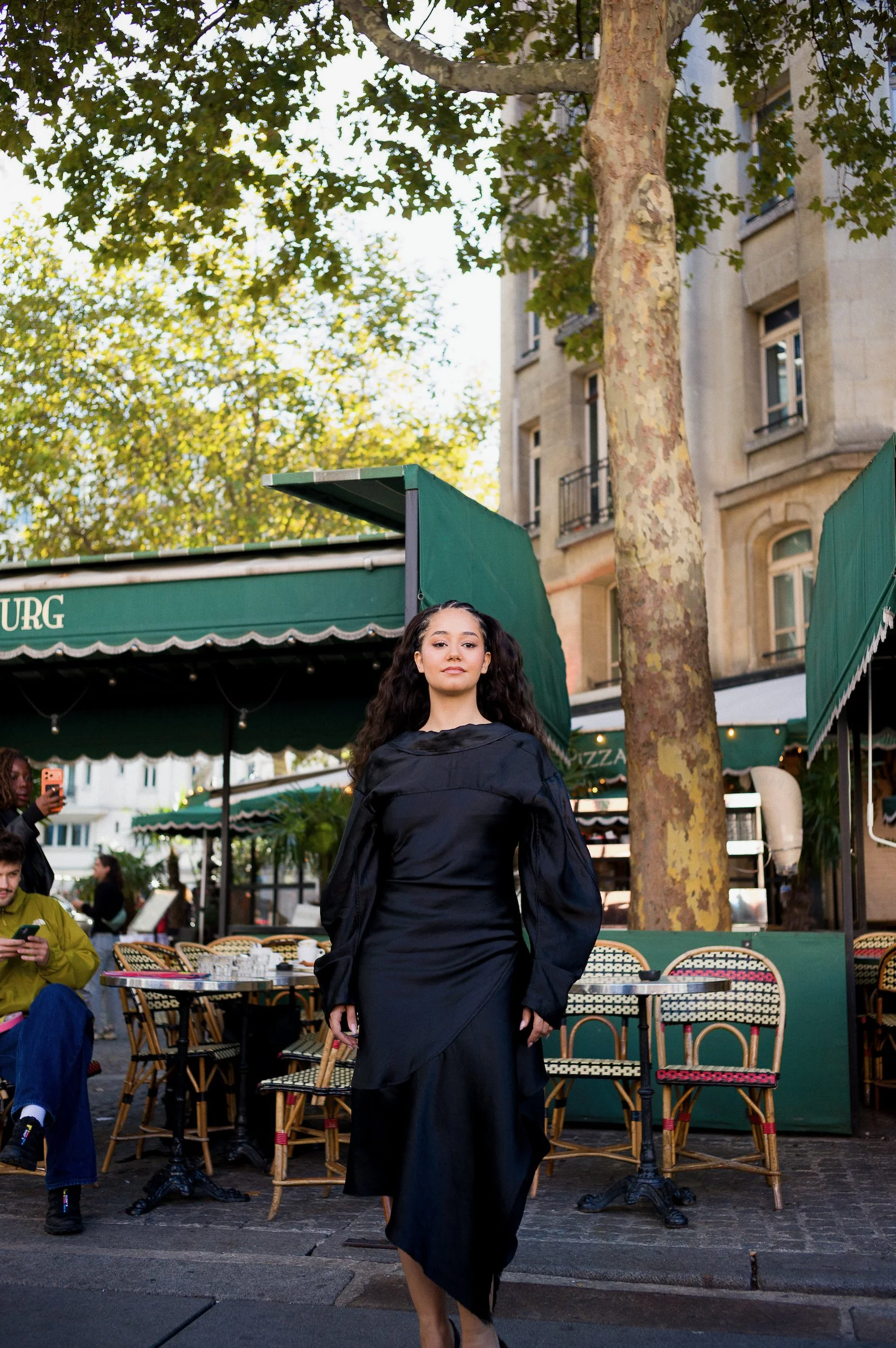 A woman with long, dark curly hair wearing a black dress and jacket walking in an outdoor cafe with rattan chairs, tables, green awnings, and trees, in a city setting with beige apartment buildings in the background.
