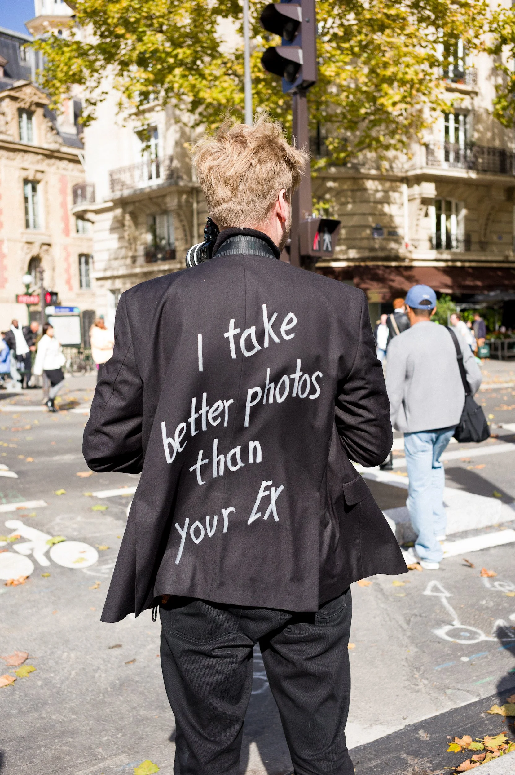At Paris Fashion Week, Back of a person wearing a black jacket with a message written in white on the back, standing on a city street with people walking around, trees with autumn leaves, and buildings in the background.