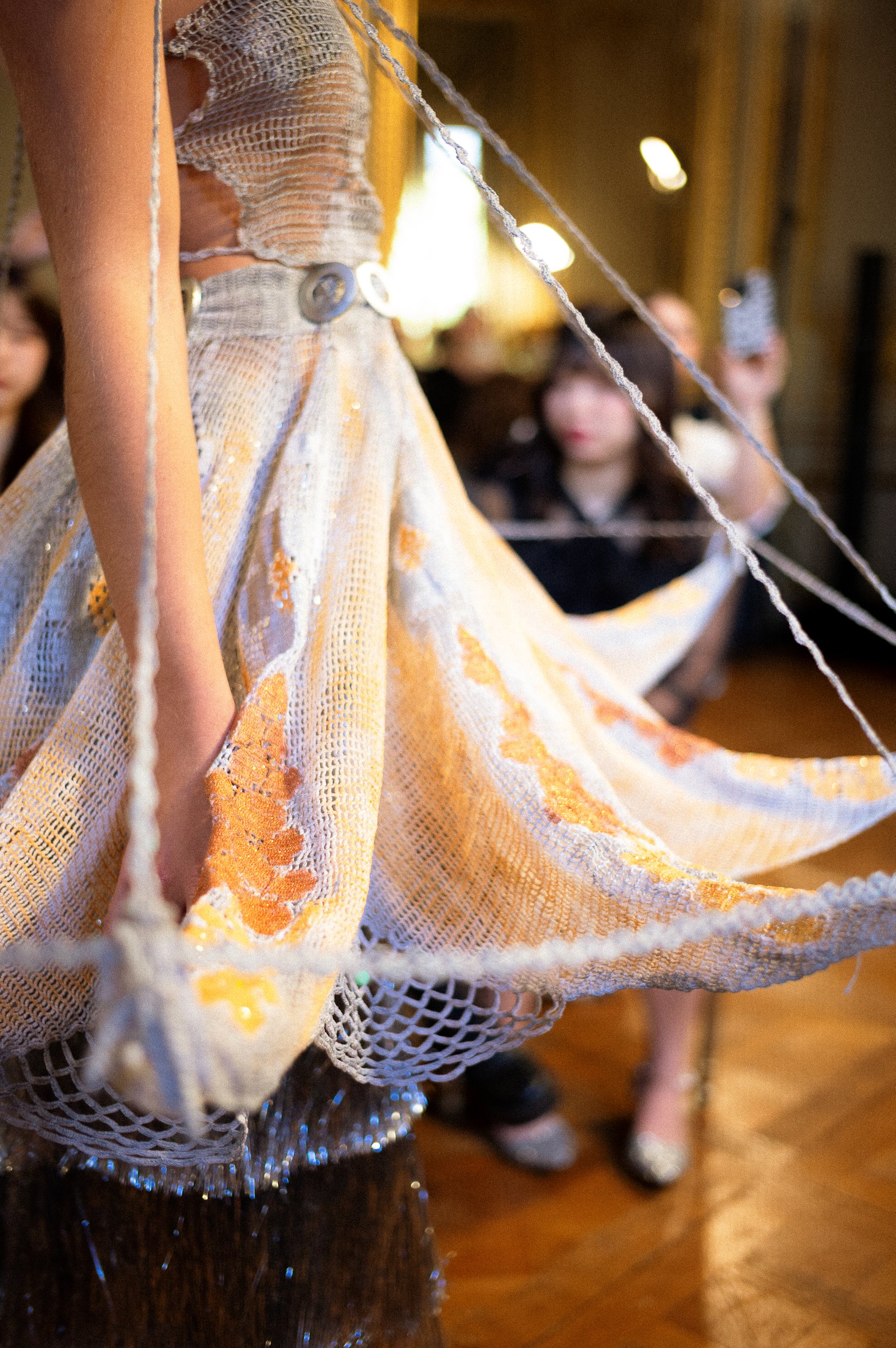 Close-up of a dress with a netted, sheer fabric in white with orange floral patterns, worn by a person with a visible arm. The dress is on a mannequin or display, with blurred background showing people, including a woman with glasses and a dark top, 