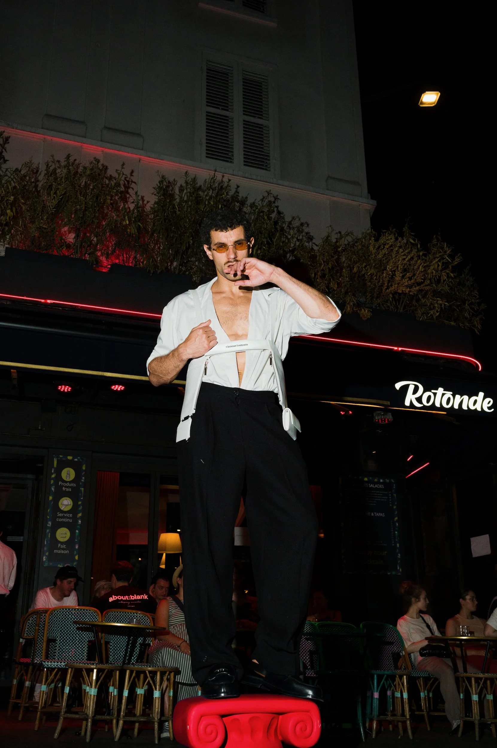 A man with curly dark hair, wearing orange-tinted glasses, a white shirt unbuttoned at the top, black pants, and shiny black shoes, standing with one foot on a red ornate stool and the other on the ground, with a building and crowd in the background 