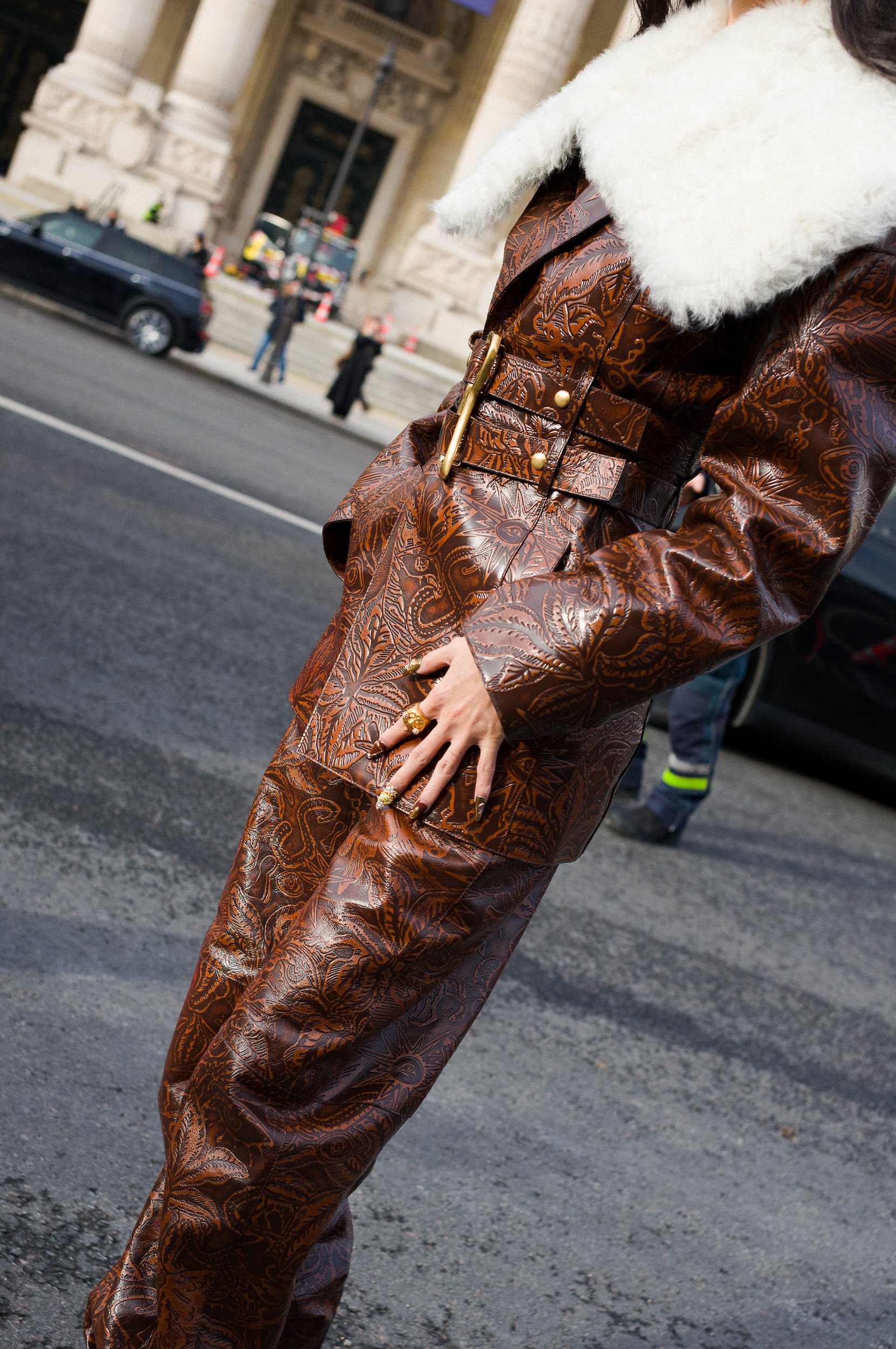 At Paris Fashion Week, Close-up of a person wearing a brown patterned leather outfit with a white fluffy collar, standing on a city street.
