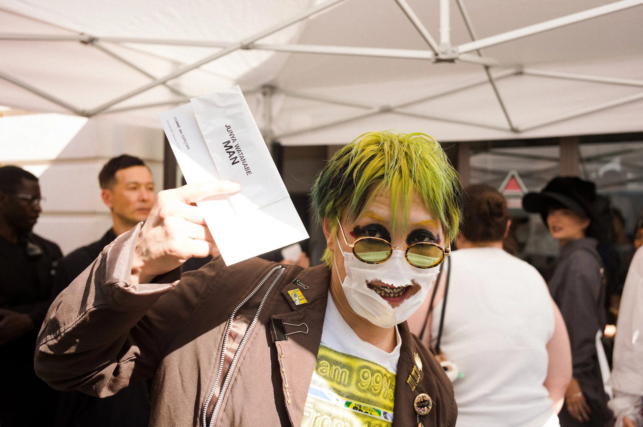 Person with bright green hair and sunglasses, wearing a face mask, holding a ticket, standing among a crowd under a white canopy.