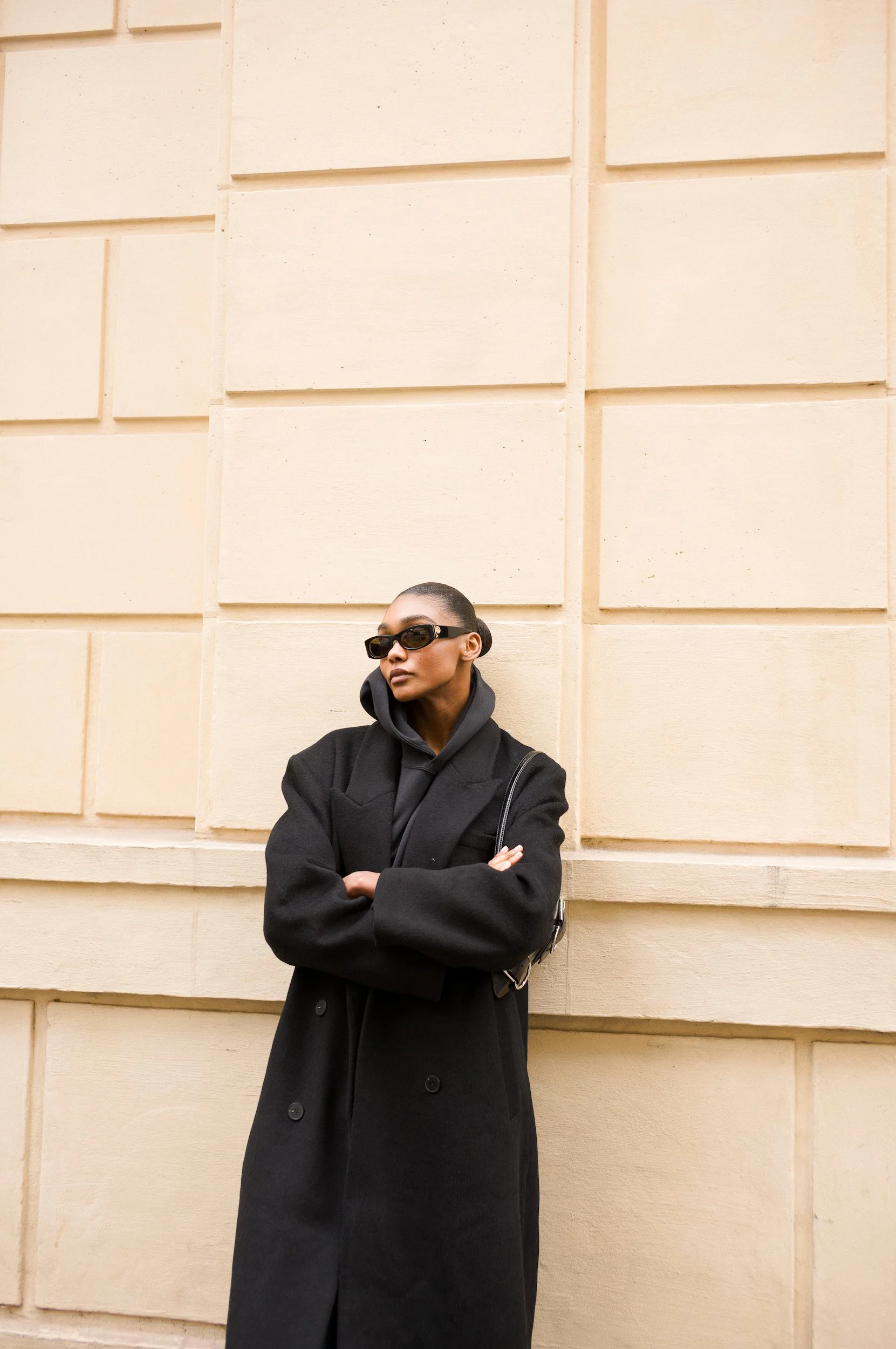 At Paris Fashion Week, A woman dressed in black with sunglasses standing in front of a beige wall with rectangular panels, arms crossed.