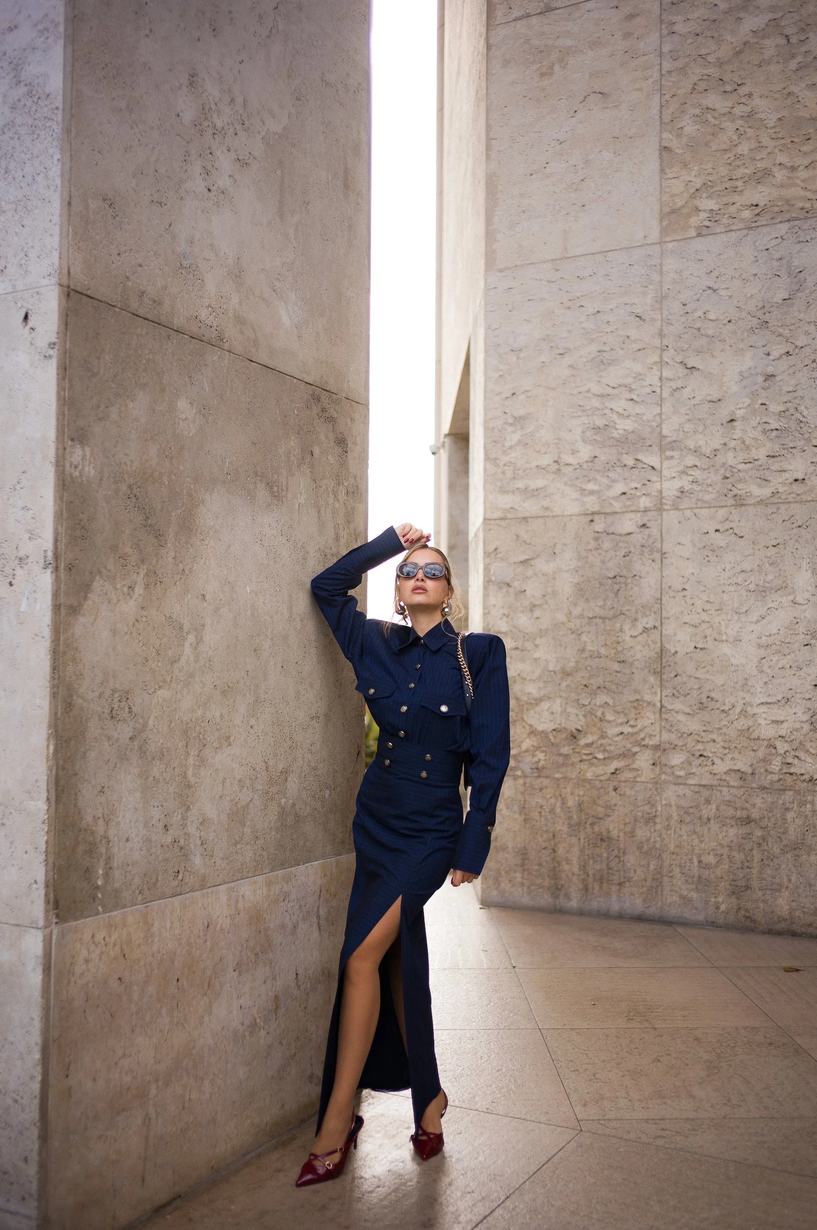 At Paris Fashion Week, A woman wearing a navy blue dress with a high slit, red high heels, large sunglasses, and jewelry, is posing outdoors against tall beige stone walls.