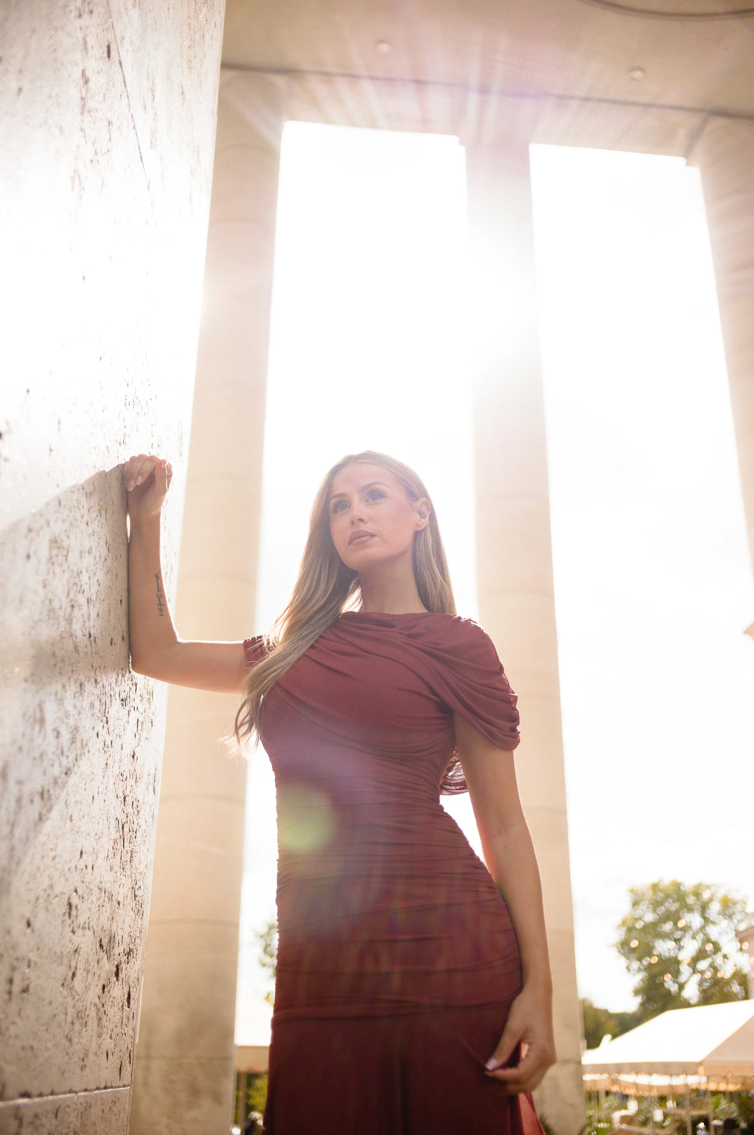At Paris Fashion Week, A woman in a fitted maroon dress stands outdoors near a stone wall with columns, with sunlight behind her, creating a backlit effect.