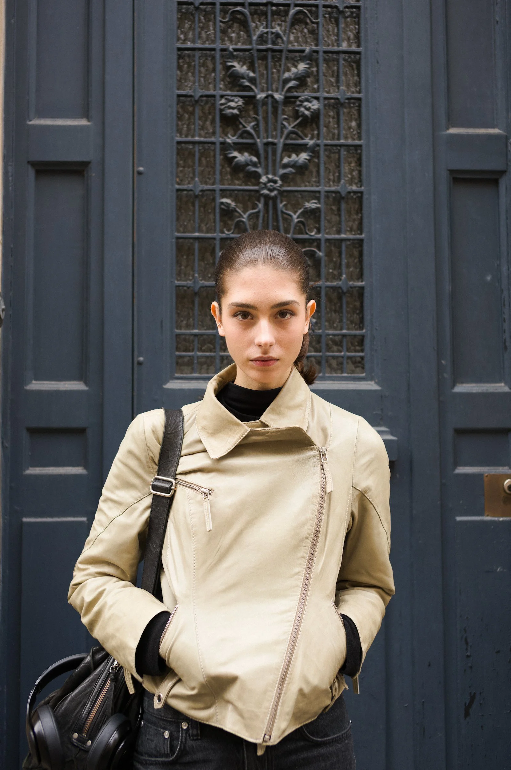 A young woman with dark brown hair in a ponytail, wearing a beige jacket, black top, and black jeans. She has a black bag over her left shoulder and holds a pair of black headphones in her left hand. She is standing in front of a large, dark blue woo