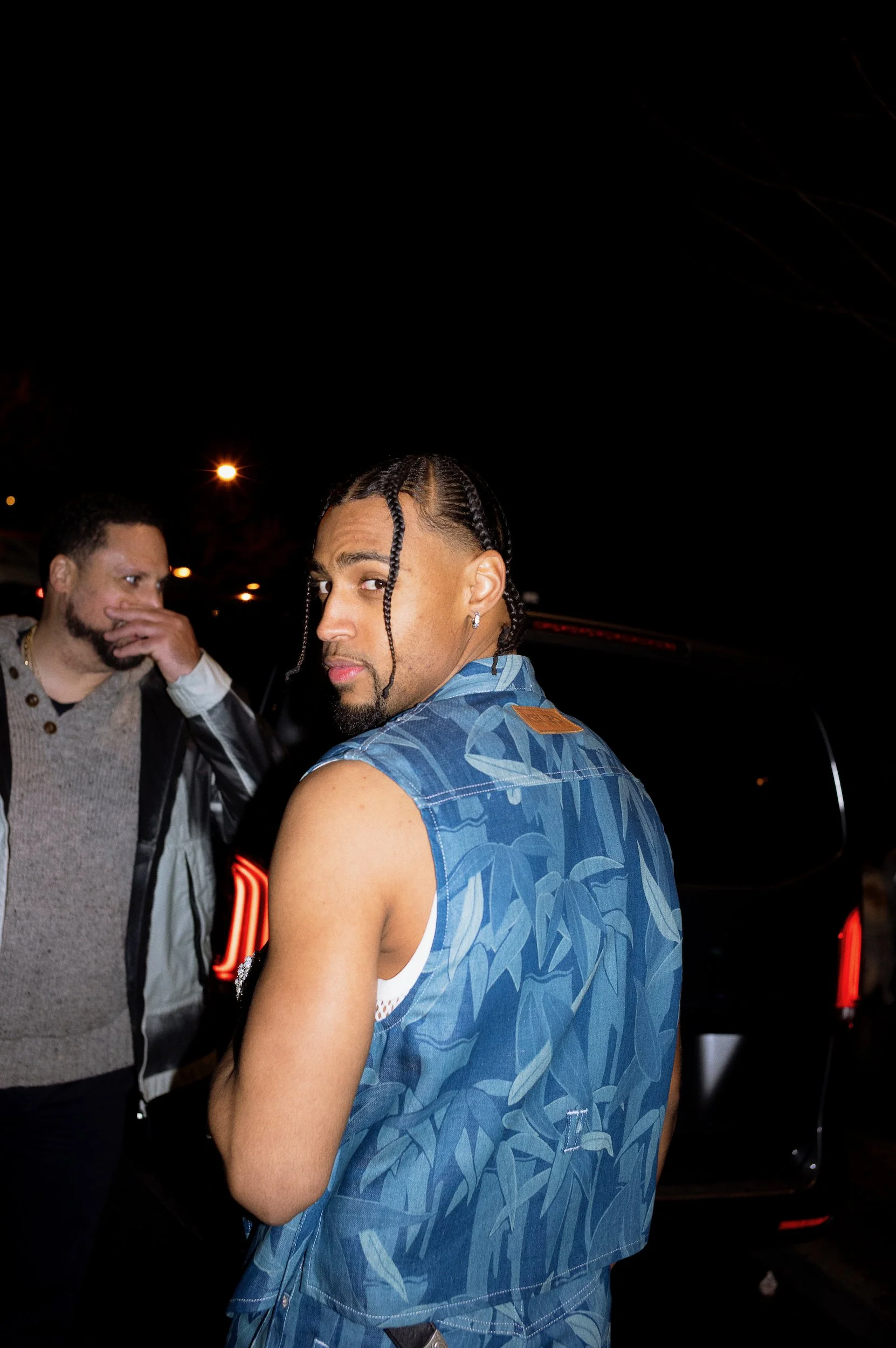 At Paris Fashion Week, A young man with braided hair and earrings wearing a blue patterned sleeveless shirt looking at the camera with a serious expression, at night with a black car in the background and a man covering his mouth.