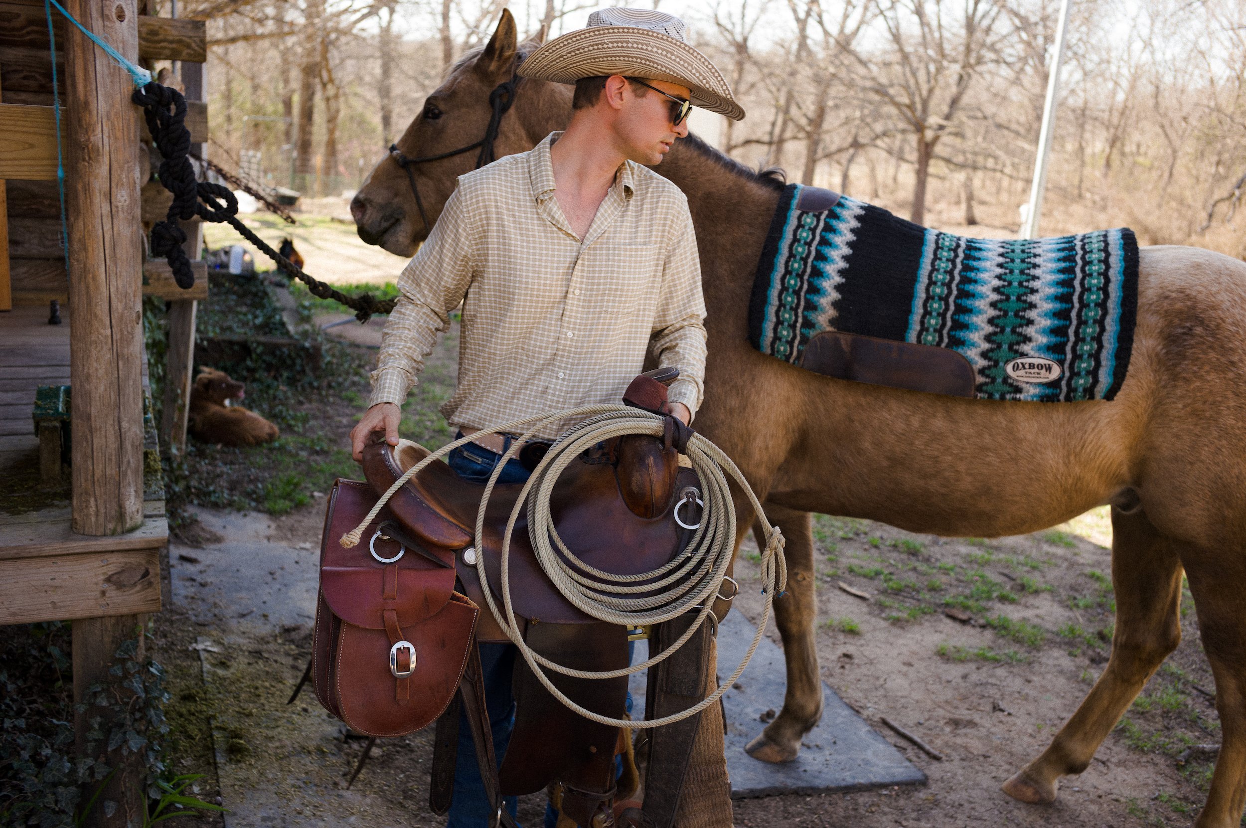Cowboy wearing a plaid shirt, sunglasses, and a wide-brimmed hat is standing beside a brown horse with a patterned blanket. The man is holding riding gear and is near a wooden structure outdoors, with trees in the background.