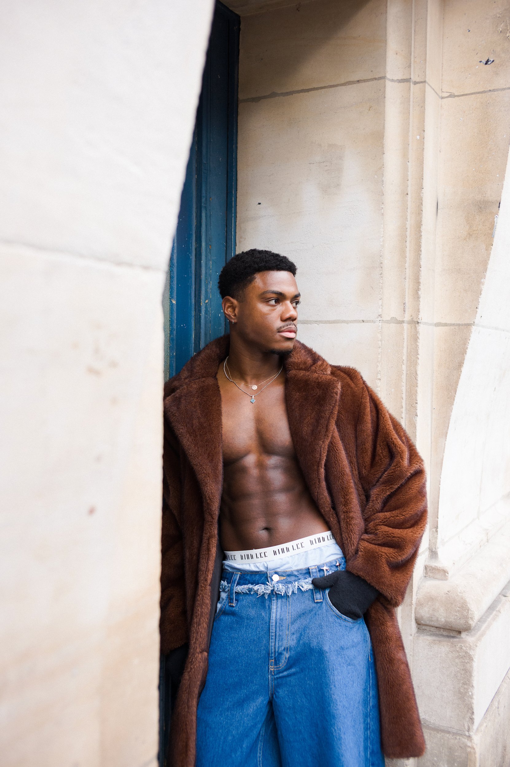 A young man with dark curly hair and medium brown skin, standing against a stone wall and doorway. He's wearing a brown fur coat, blue distressed jeans, a white underwear waistband visible, and multiple layered necklaces. His expression is serious, a