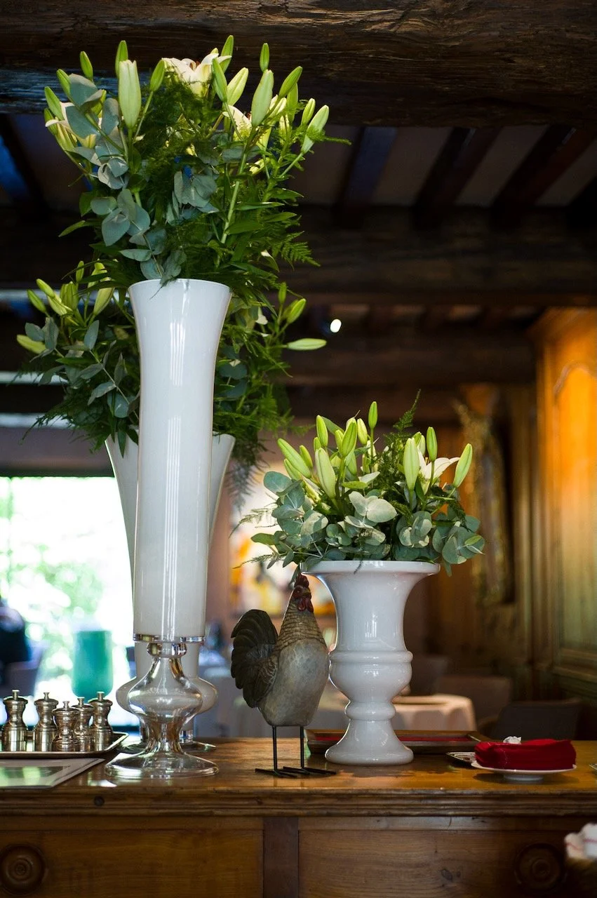 Two white vases filled with white lilies and greenery on a wooden table, with a decorative rooster sculpture, in a cozy restaurant interior.
