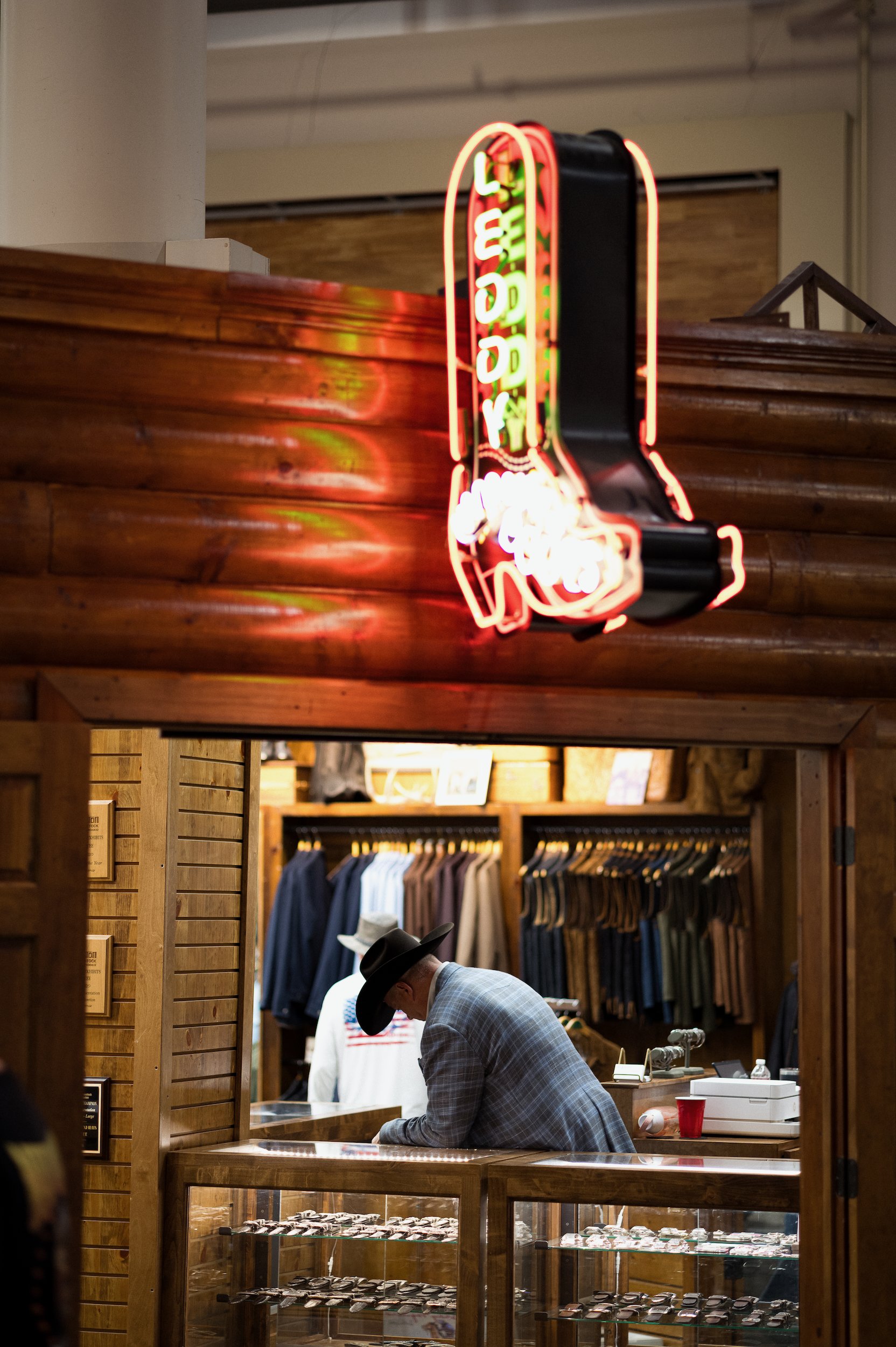 A man wearing a black cowboy hat and a gray checkered blazer is shopping in a store selling jewelry. The store has wooden walls. In the background, there are clothing racks with shirts, and ML Leddys neon sign hangs from the wall.