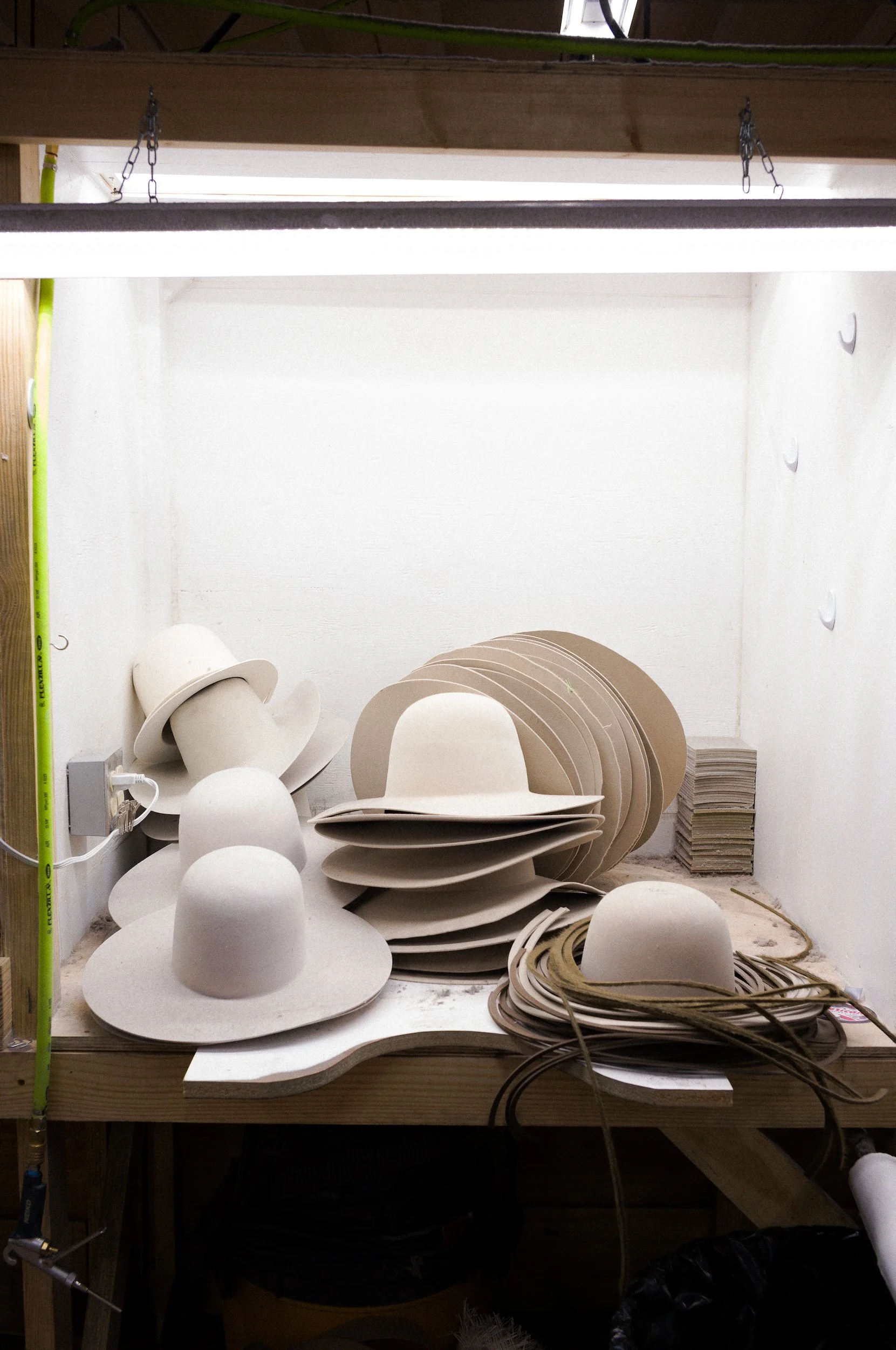 Workshop in American Hat Company with various white cowboy hats, stacked and arranged on a work table, surrounded by paper rolls, wires, and tools.