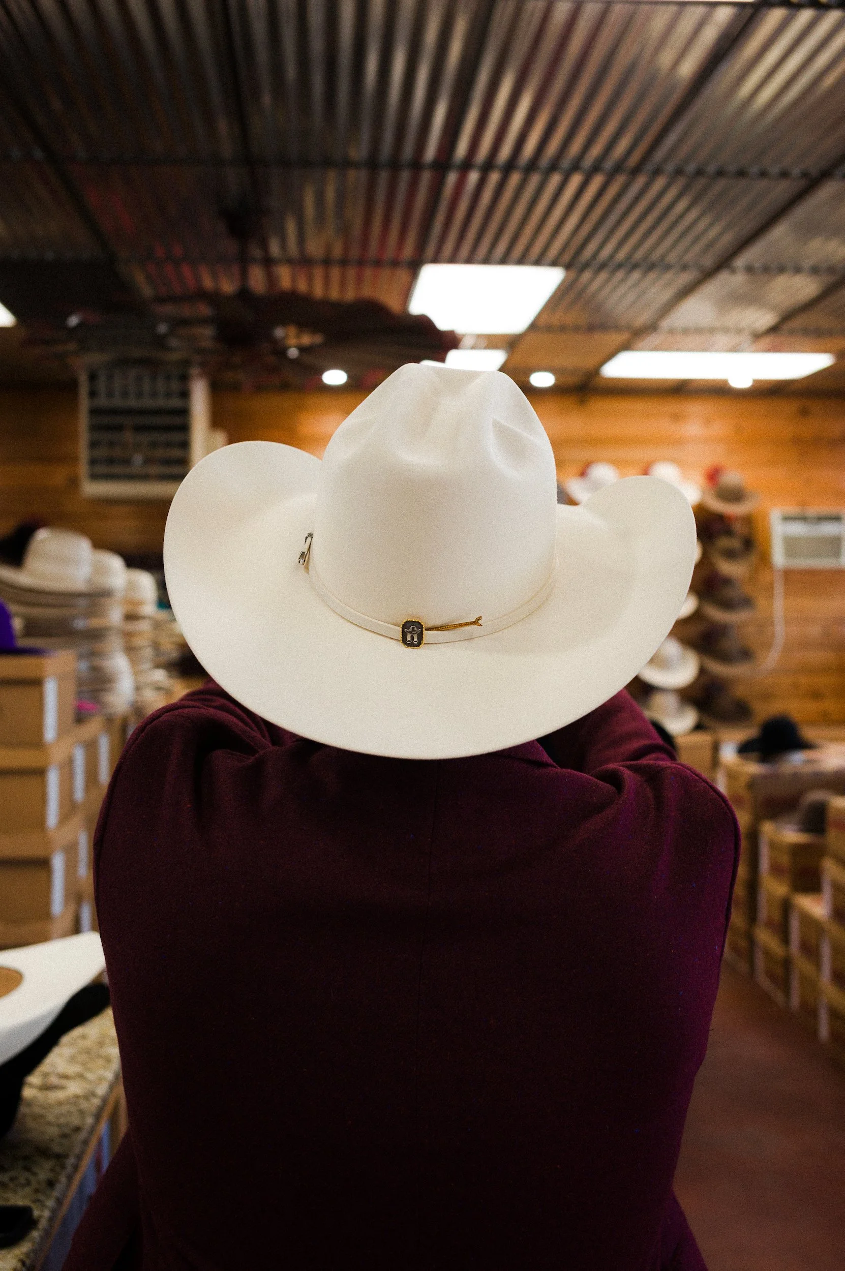 White cowboy hat with decorative band on person's head, store interior with wooden walls, various hats on shelves.