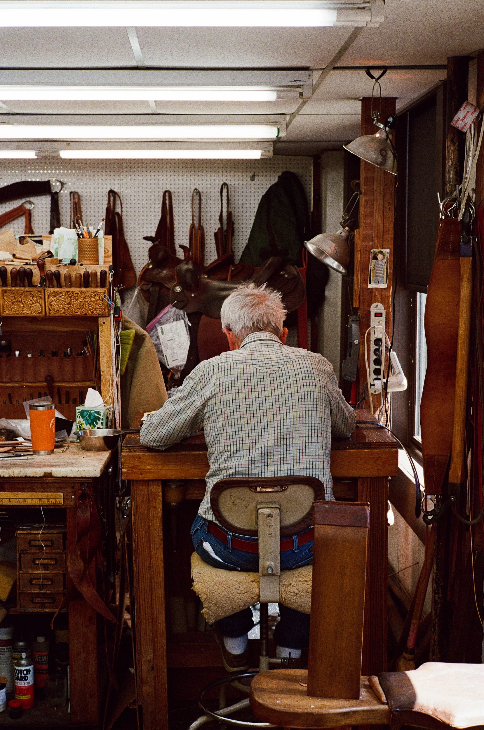 In ML Leddys, Man with gray hair wearing plaid shirt sitting on a stool inside a leather workshop at ML Leddys.