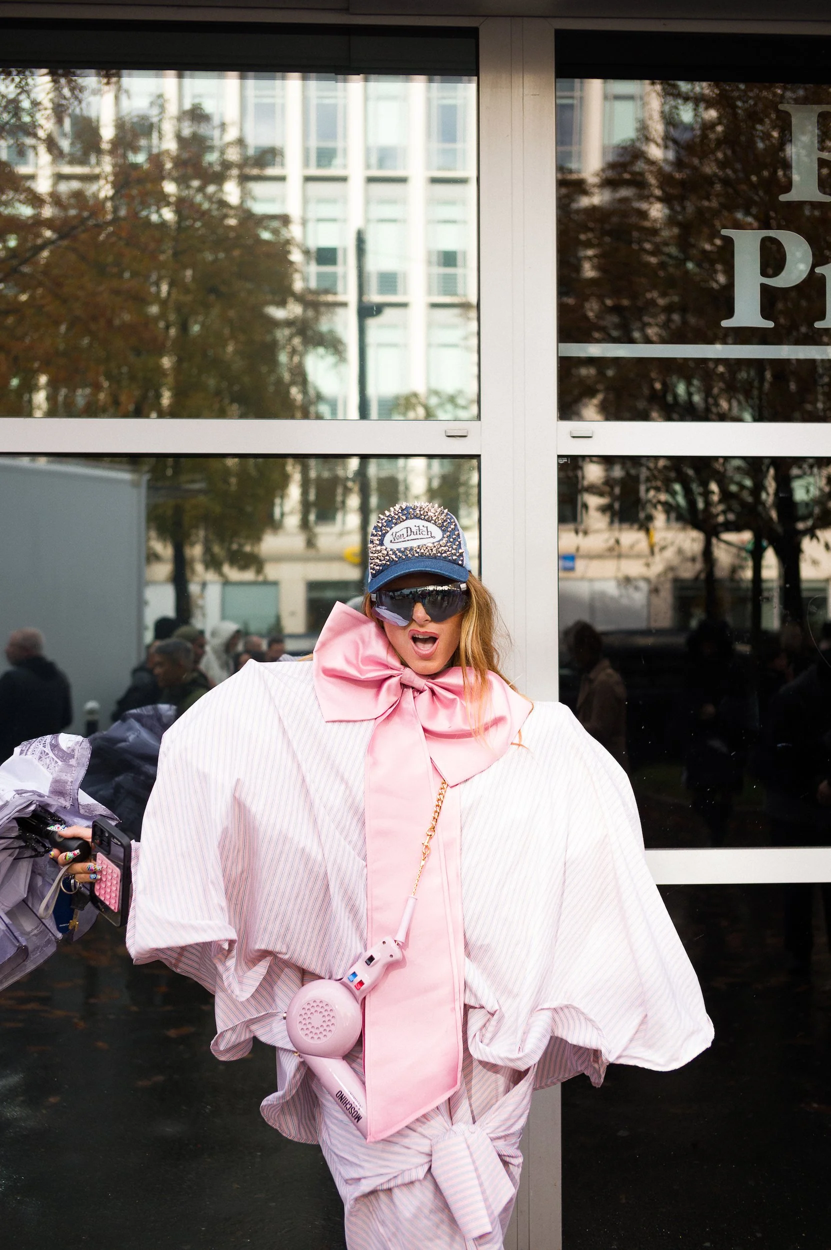 At Paris Fashion Week, Person wearing a striped oversized jacket, pink hoodie, large pink bow necklace, and a Von Dutch baseball cap, holding a pink megaphone with a pink headset, walking past a building with reflective glass windows.