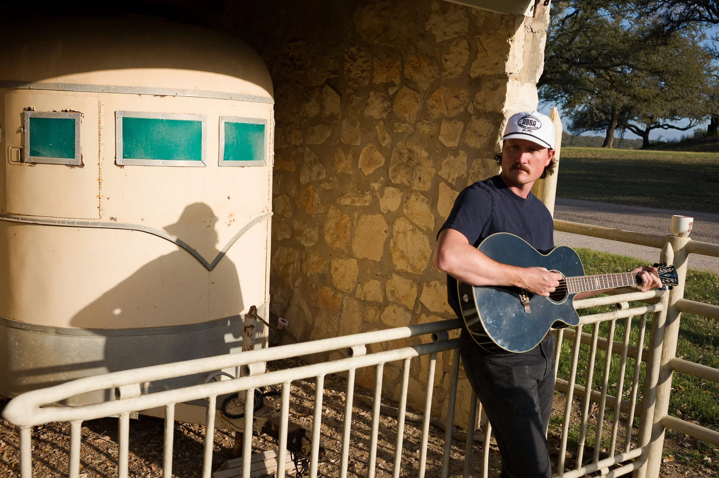 A man with a mustache wearing a white cap and a dark T-shirt is playing a blue acoustic guitar outdoors near a stone building with green windows and a wooden railing. The background includes trees and a clear sky.