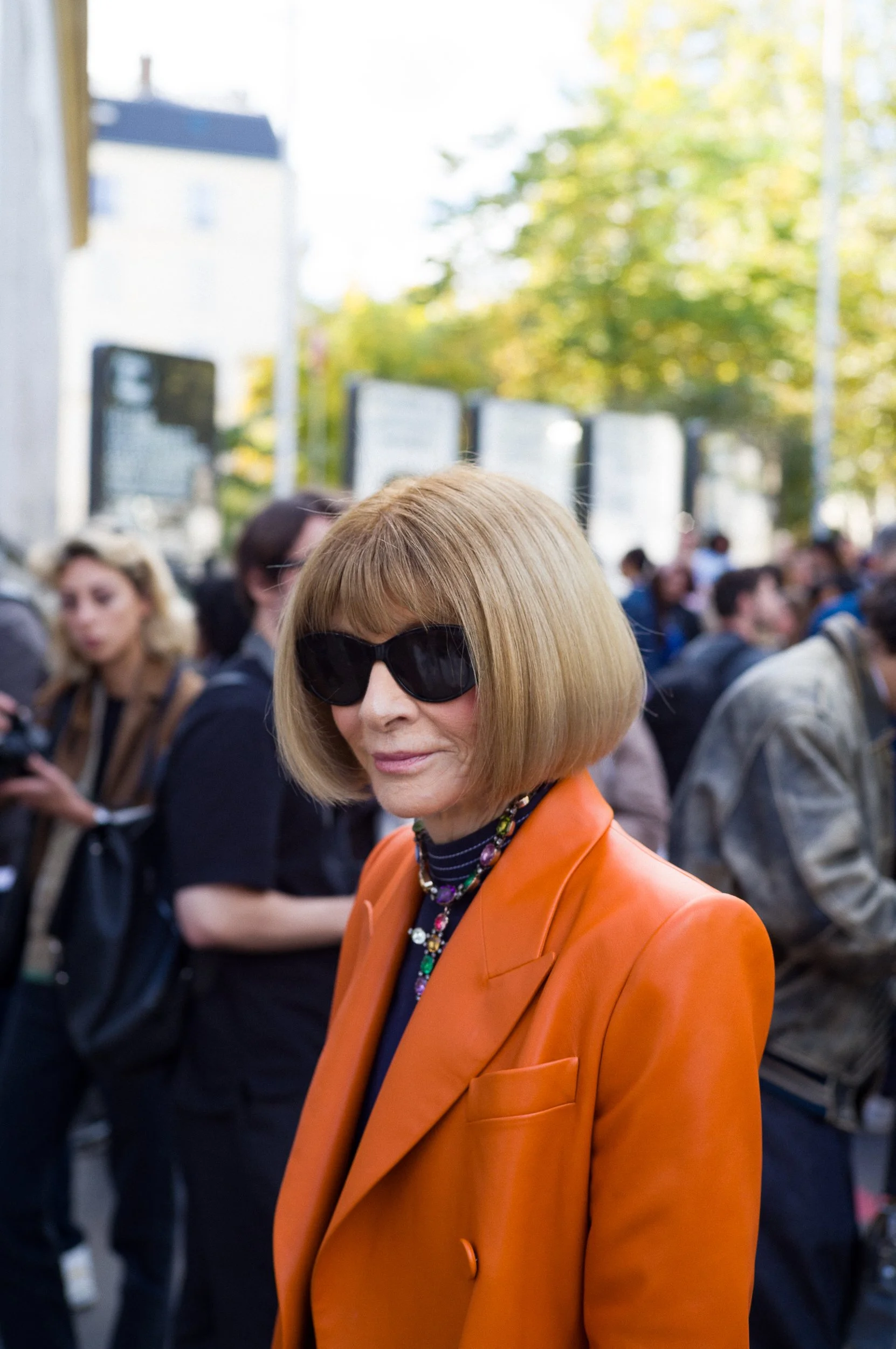 Anna Wintour with chin-length blonde hair wearing large black sunglasses, an orange blazer, and a colorful beaded necklace. She is outdoors with a crowd of people and trees with green and yellow leaves in the background.