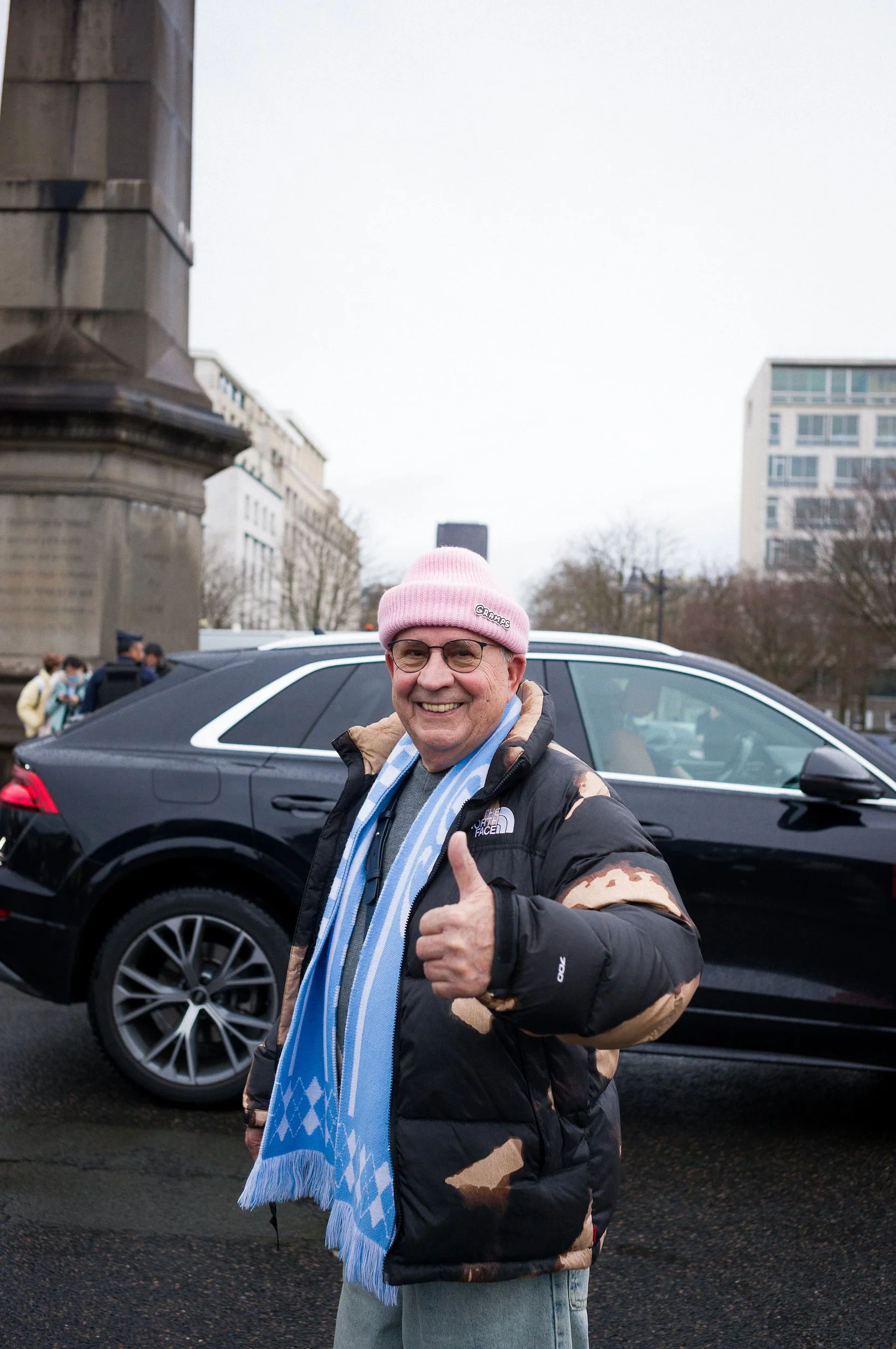 At Paris Fashion Week, A smiling man wearing a pink beanie, glasses, a black and beige patterned jacket, and a blue and white scarf giving a thumbs-up gesture standing in front of a black car, with a city street and buildings in the background.
