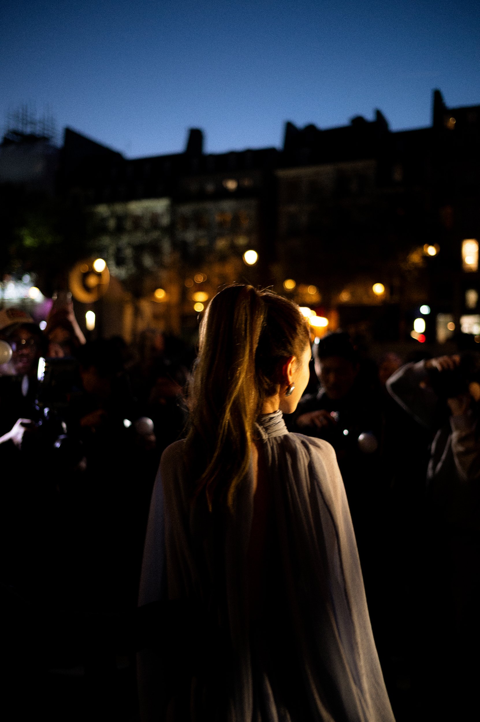 At Paris Fashion Week, A woman with long blonde hair standing at night in a crowd, with city buildings and streetlights in the background.