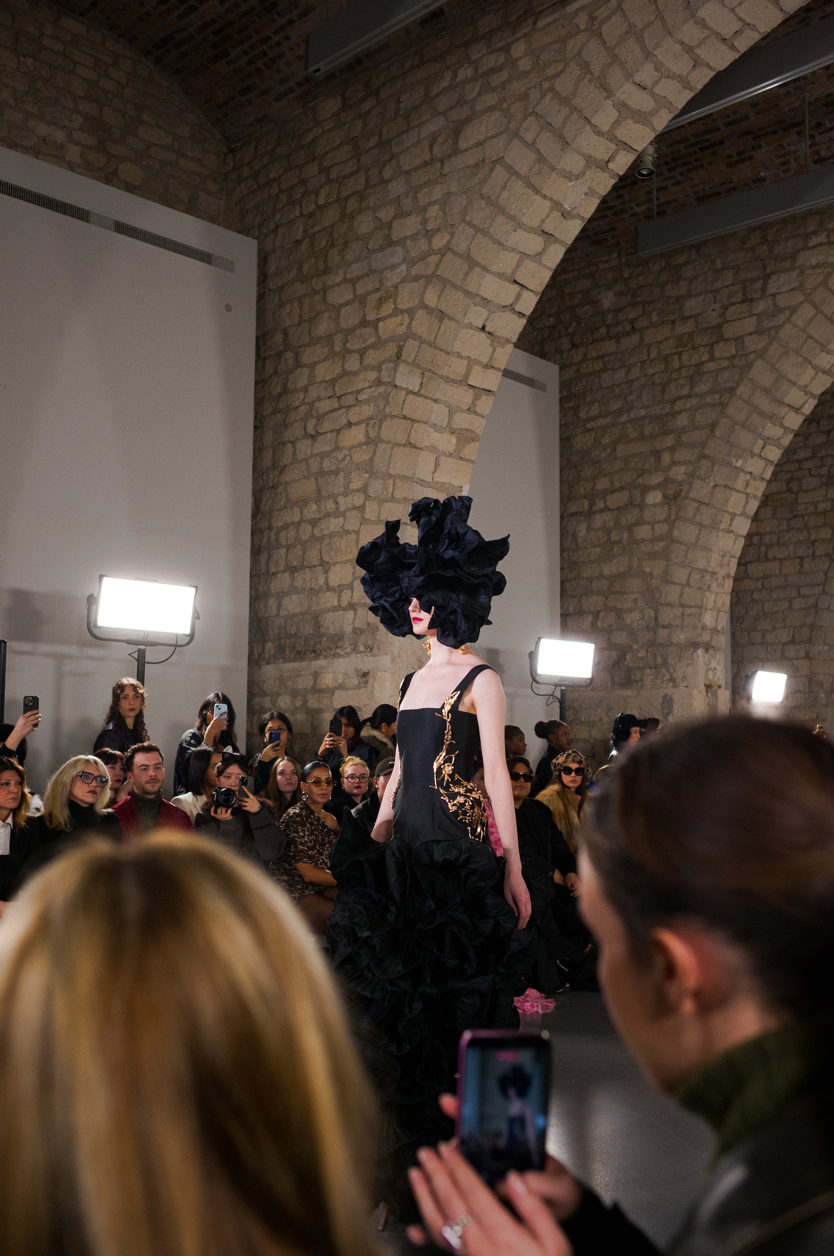 A runway model with an elaborate black floral headpiece and a black dress with gold embroidery is walking down the runway at a fashion show. The venue features exposed brick arches and is crowded with seated and standing audience members, some captur