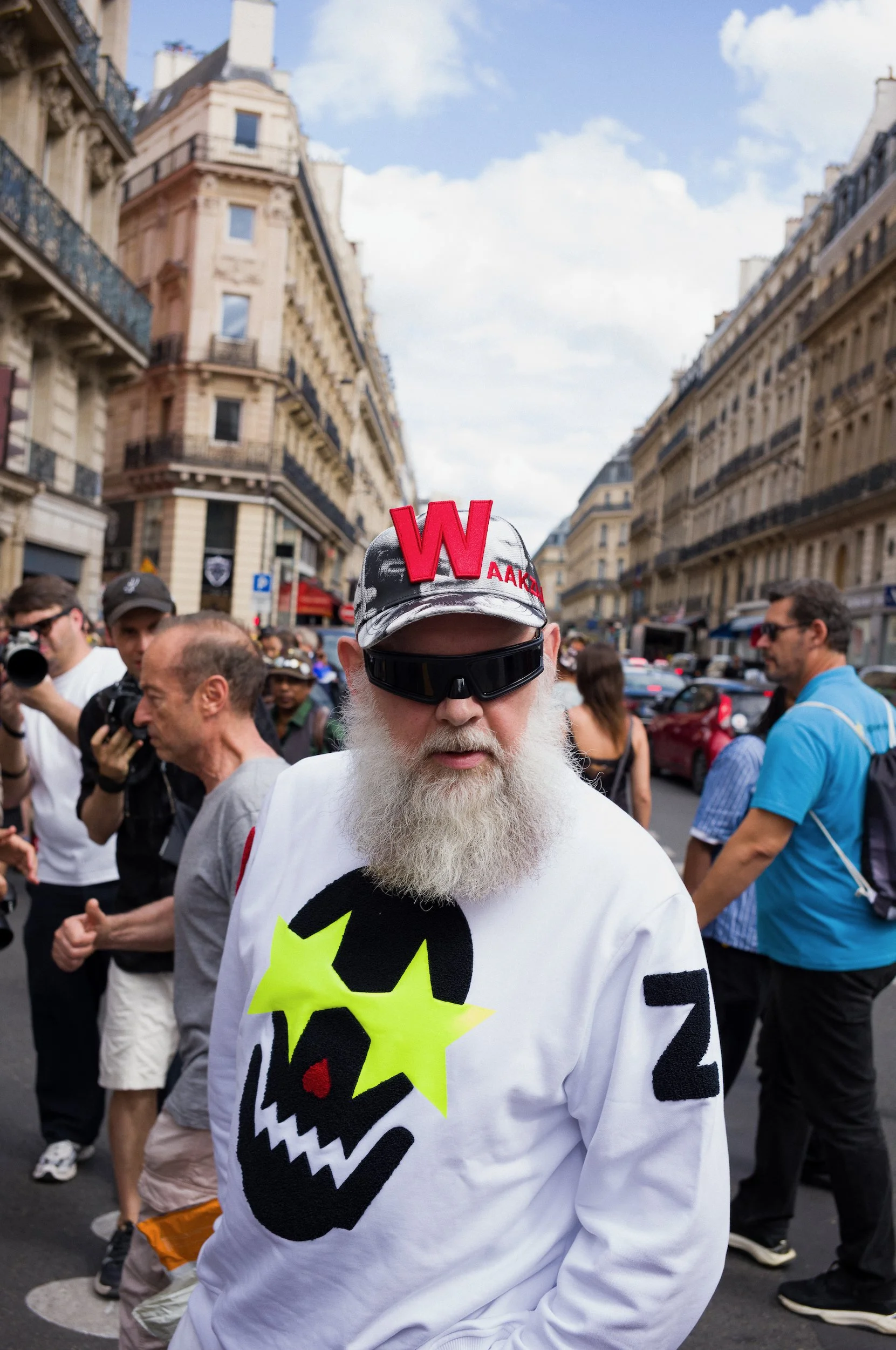 Walter Van Beirendonck wearing a cap with a red letter W and black sunglasses walks through a crowd on a city street during a protest or demonstration. The man is wearing a white shirt with colorful graphics, including a skull and the number 22.