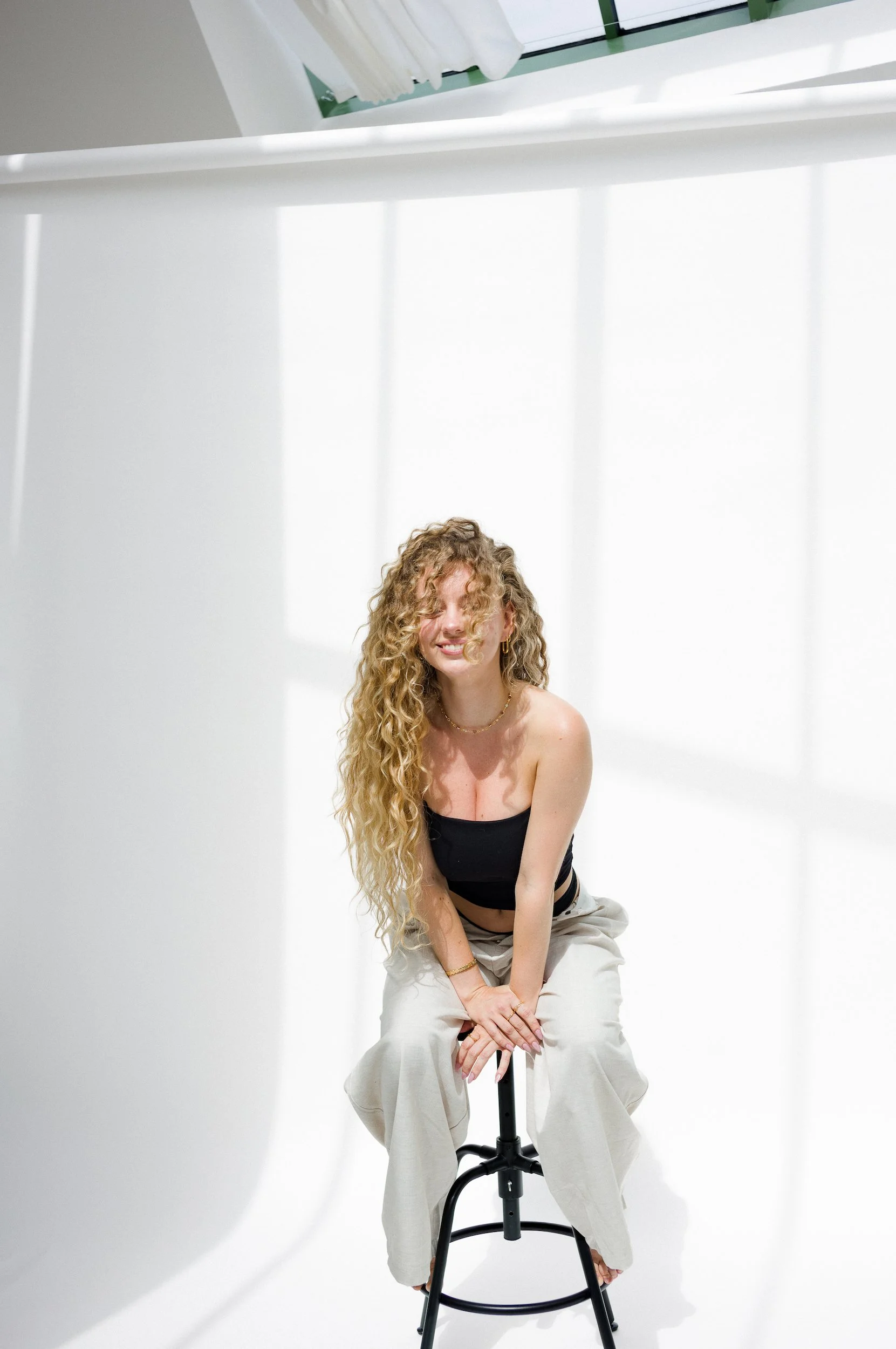 A woman with long curly hair sitting on a stool in a white studio with a bright background, smiling at the camera.