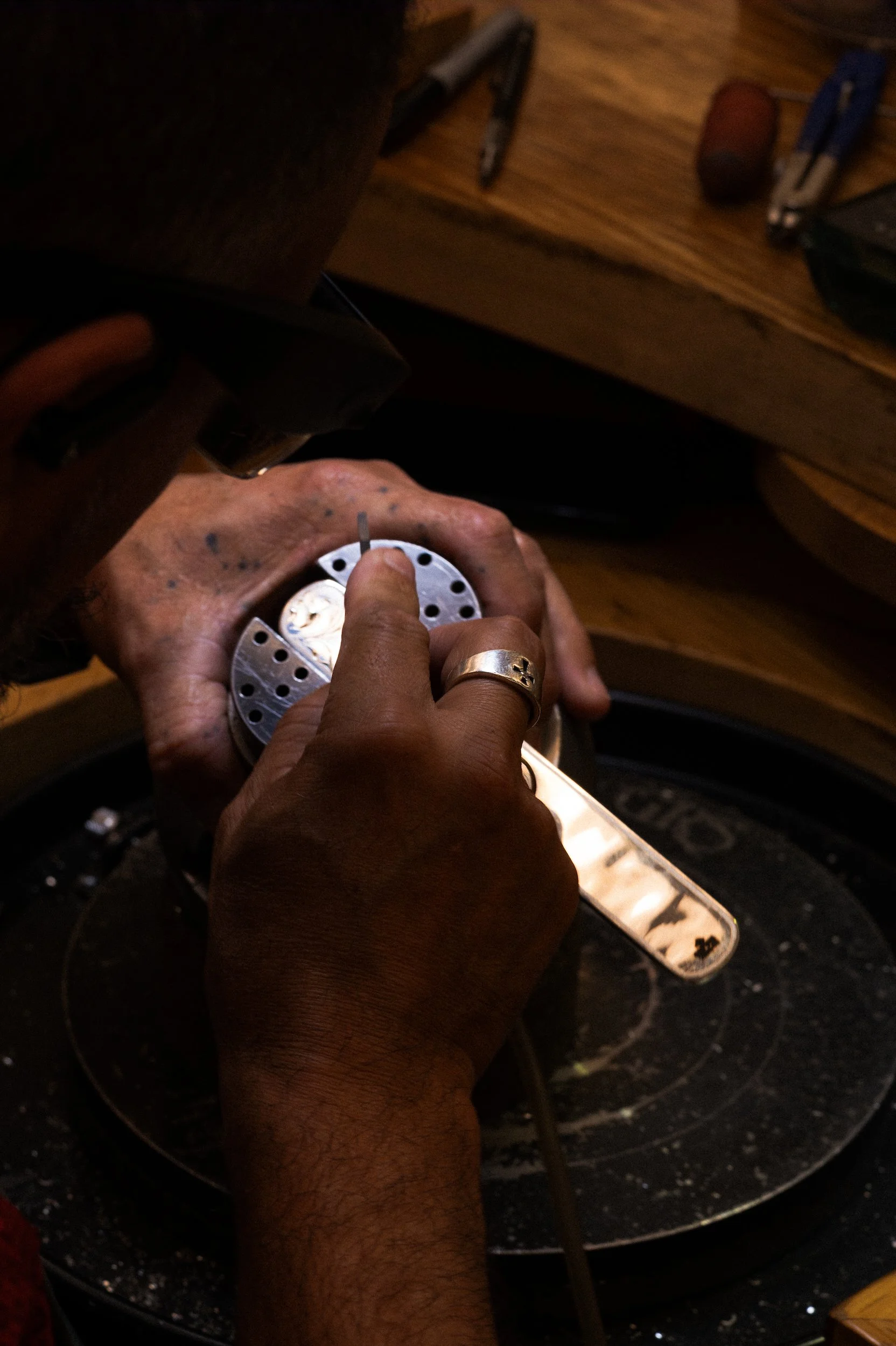 A person working on a piece of silver with a small drill or polishing tool, surrounded by various tools on a wooden workspace.