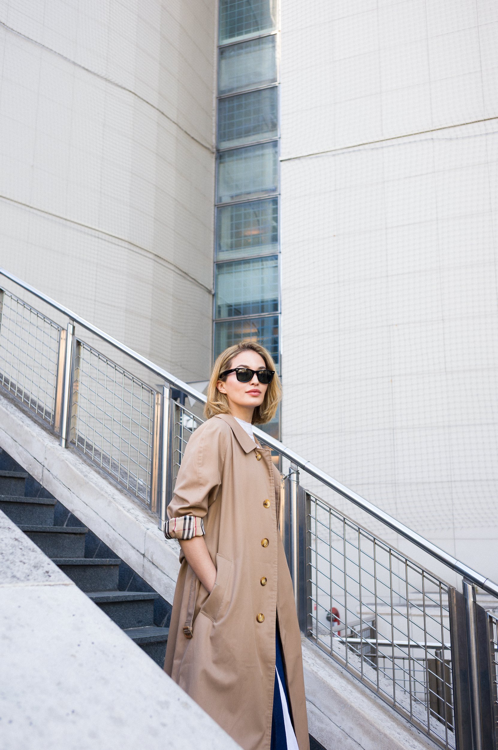 At Paris Fashion Week, A woman wearing a beige trench coat and sunglasses standing on an outdoor staircase with modern building in background.