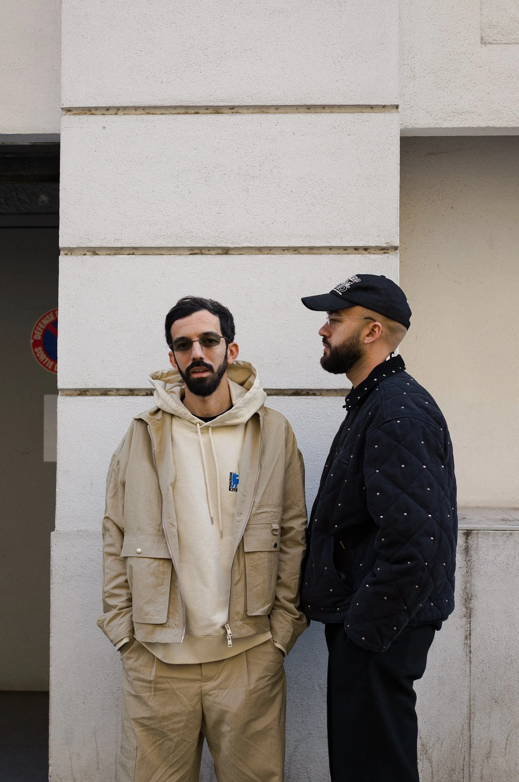 Two men standing against a white wall with horizontal lines. One man facing the camera has dark hair, glasses, a beard, and wears a beige hoodie and beige pants. The other man, in profile, has a beard, glasses, a black cap, and a black jacket with a 