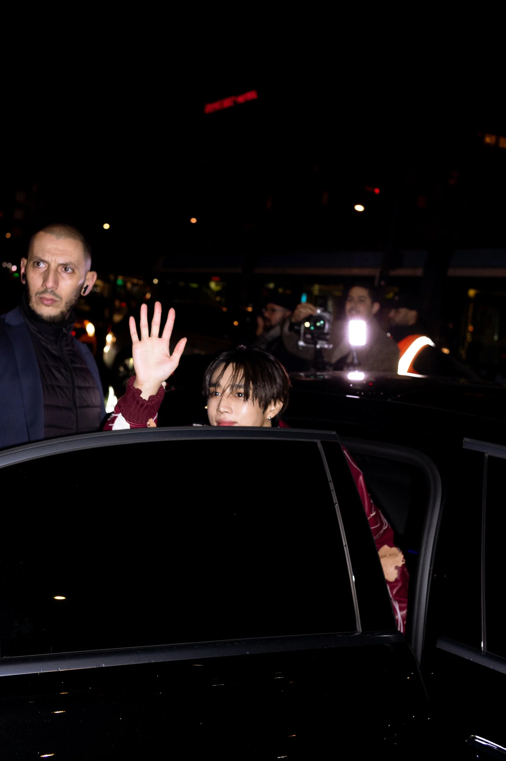 People are gathered outside at night, one person is in a car with the door open, and there are others with a camera and a flash in the background. One individual is raising a hand.