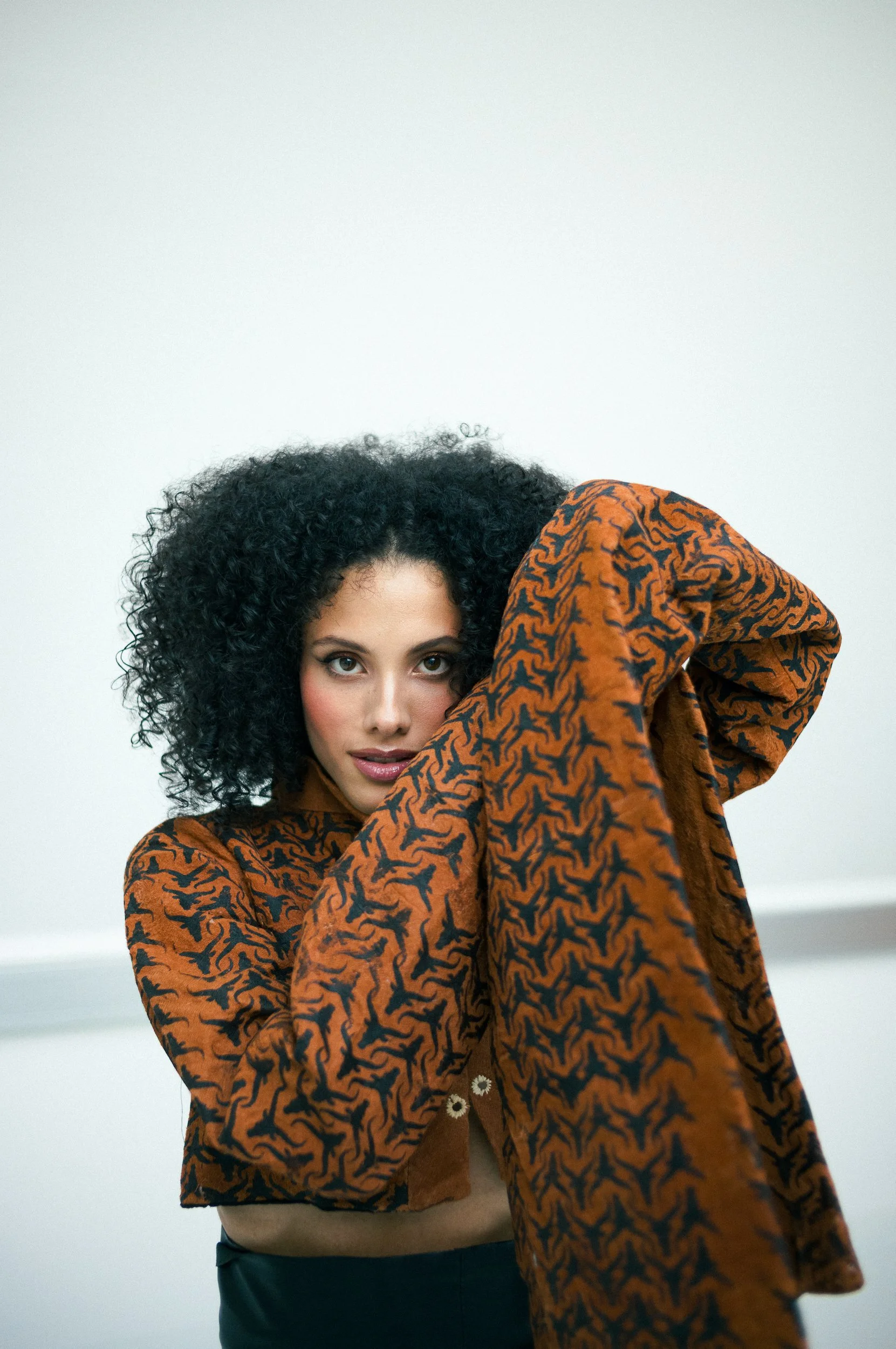 A woman with curly black hair wearing a brown and black patterned jacket, standing against a plain white background.