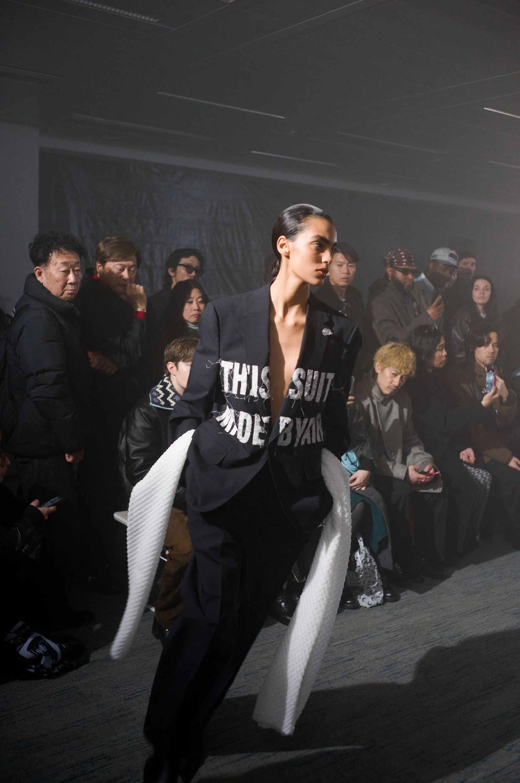 At Paris Fashion Week, Fashion runway model walking with a white textured coat draped over her shoulders, wearing a black outfit with text on it, and audience members seated and standing watching the event.