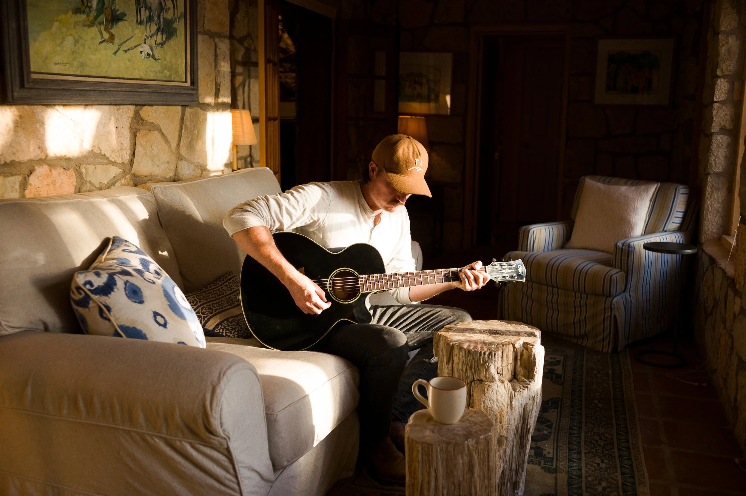A man wearing a beige cap and a white shirt is sitting on a couch playing an acoustic guitar. The room has stone walls, framed artwork, a wooden side table, and a cup on a wooden stump in front of him. Soft warm lighting creates a cozy atmosphere.