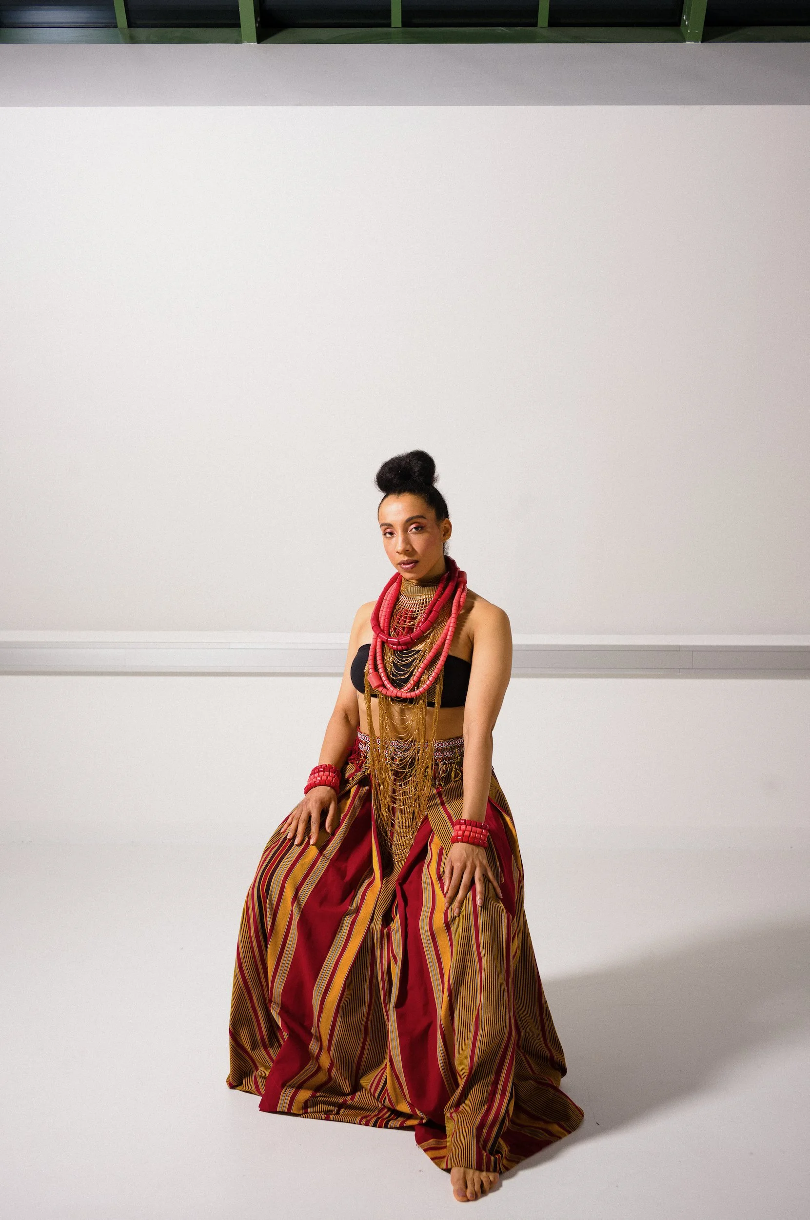 A woman of African descent in traditional attire with layered red, yellow, and black striped fabric, multiple beaded necklaces, and red jewelry, seated against a plain white background.