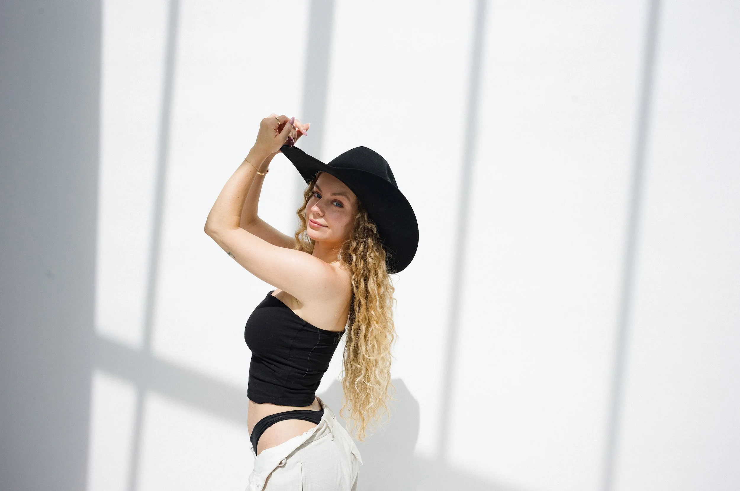A woman with long curly blonde hair wearing a large black sunhat, black strapless top, and white pants with the waistband pulled down, standing against a white wall with shadows.