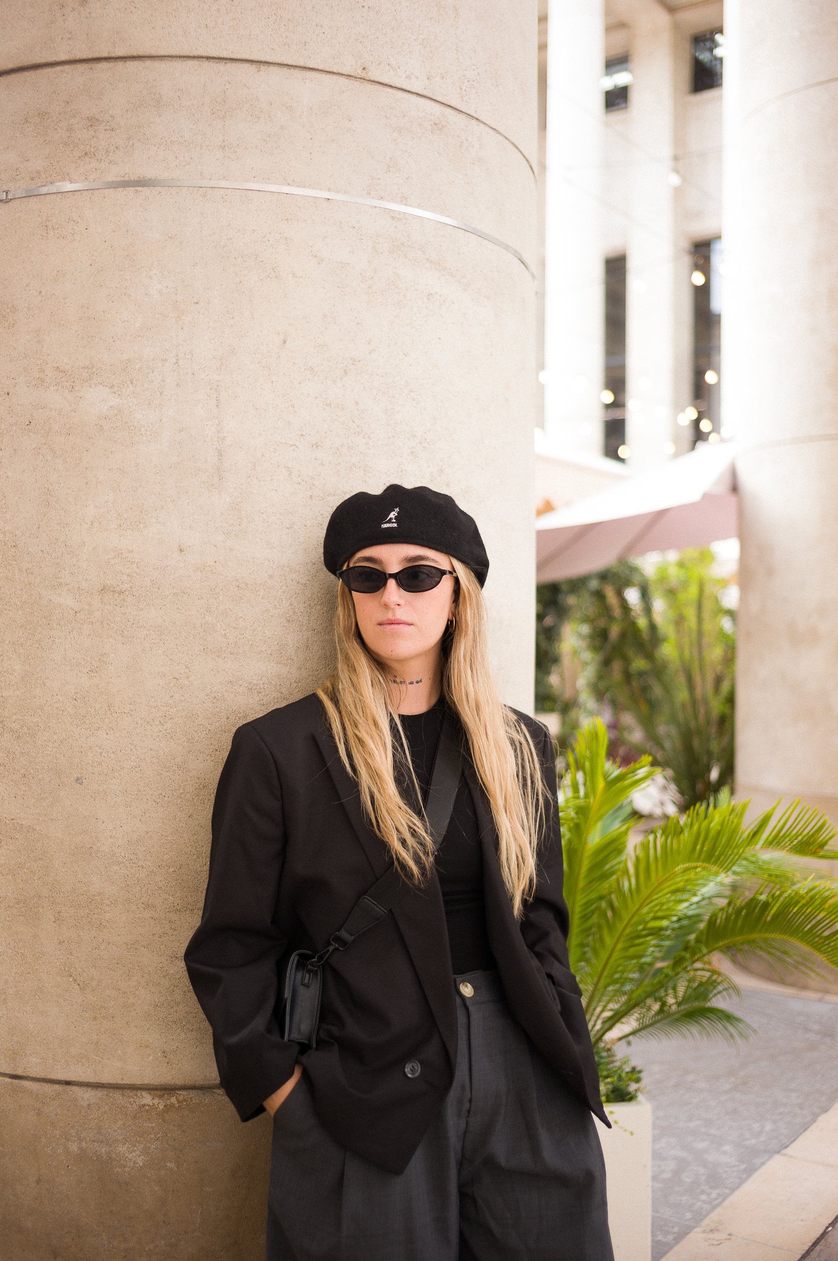 At Paris Fashion Week, A woman with long blonde hair standing outdoors near a curved concrete wall and green plants, wearing a black beret, sunglasses, a black blazer, and dark high-waisted pants.
