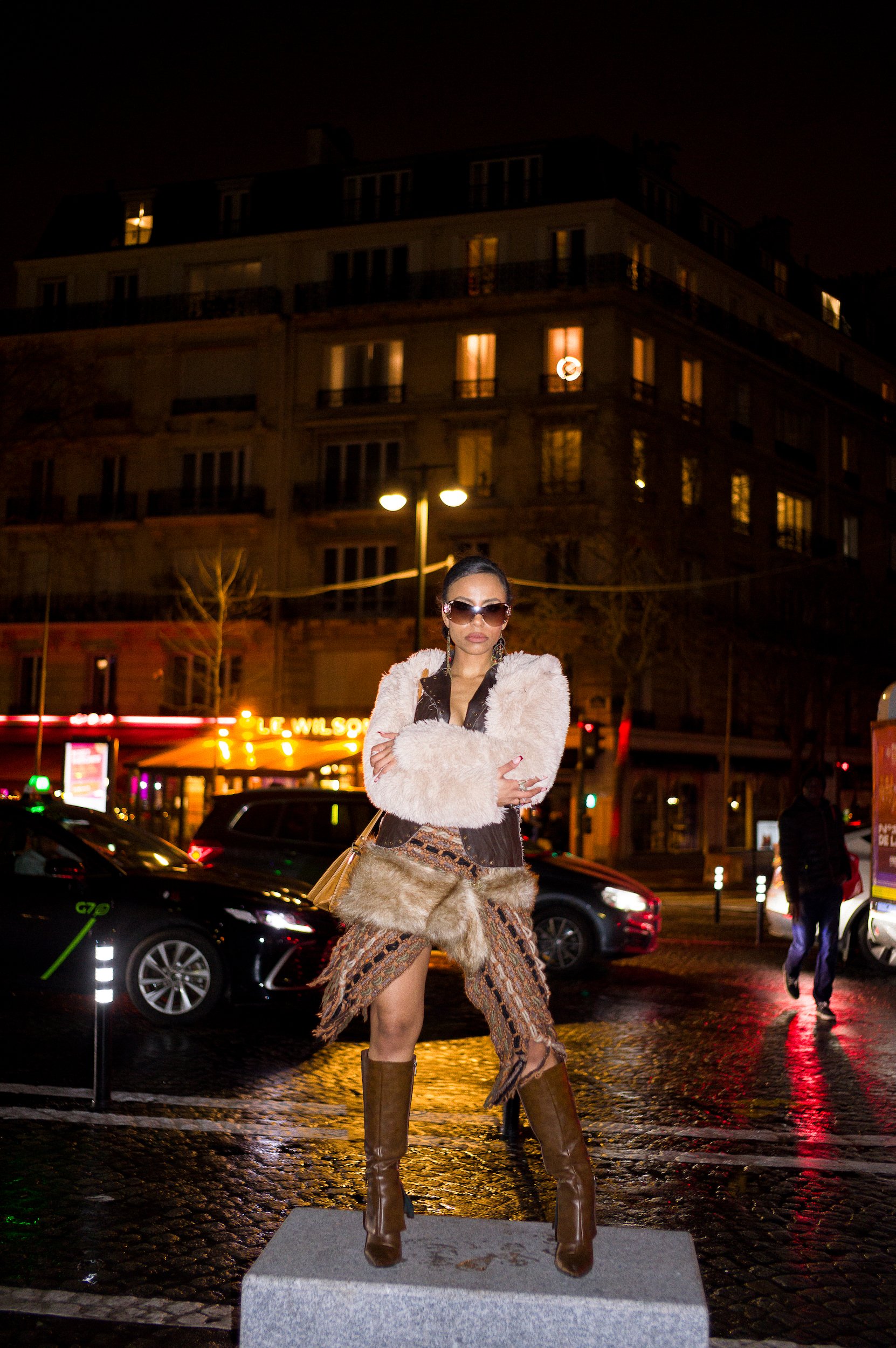 At Paris Fashion Week, A woman wearing sunglasses, a furry coat, patterned skirt, and tall boots standing on a gray pedestal at night with city lights and buildings in the background.