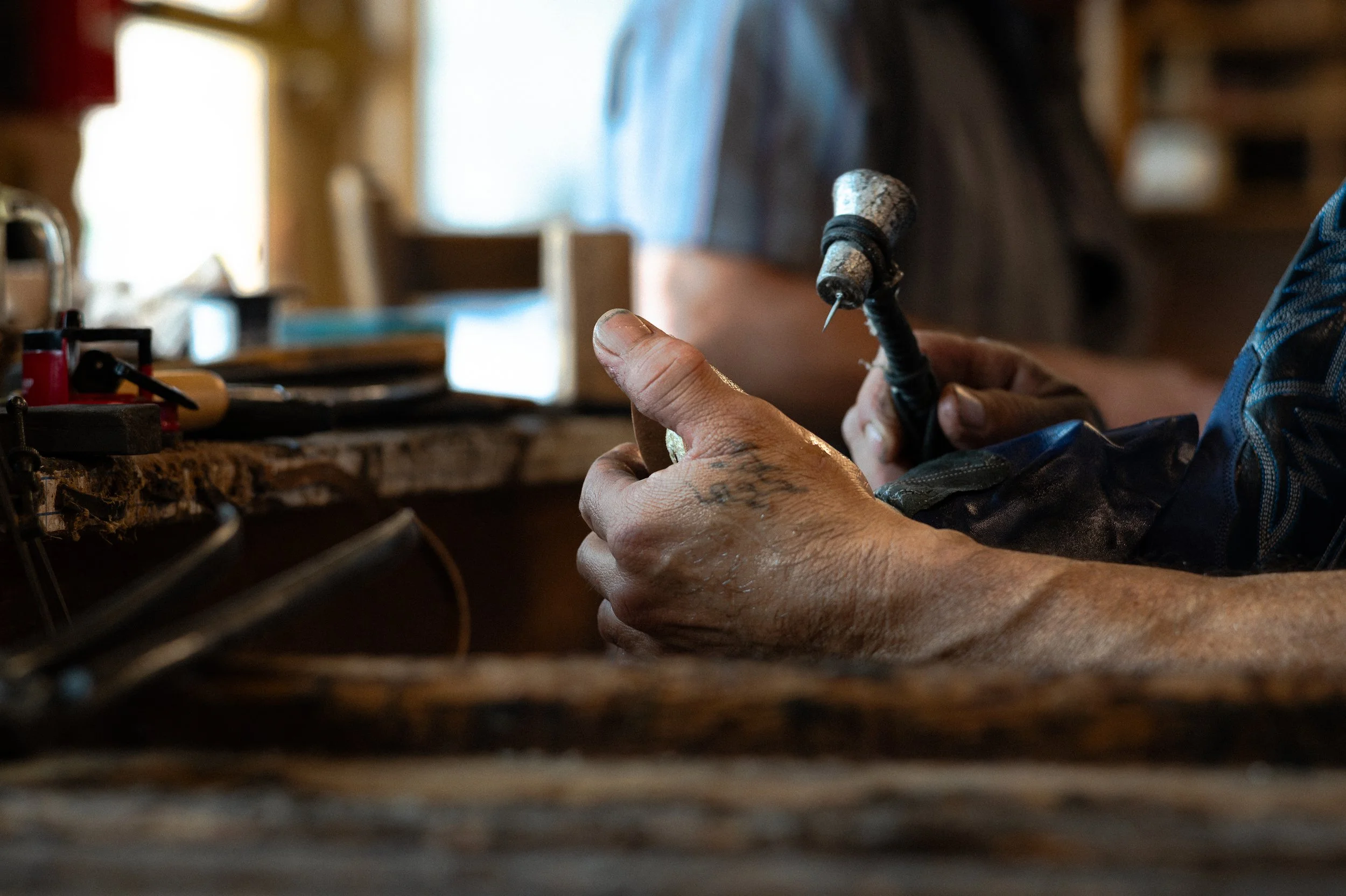 Close-up of a person's hands holding a small hammer while working on a cowboy boot at a cluttered workbench in a workshop or garage.