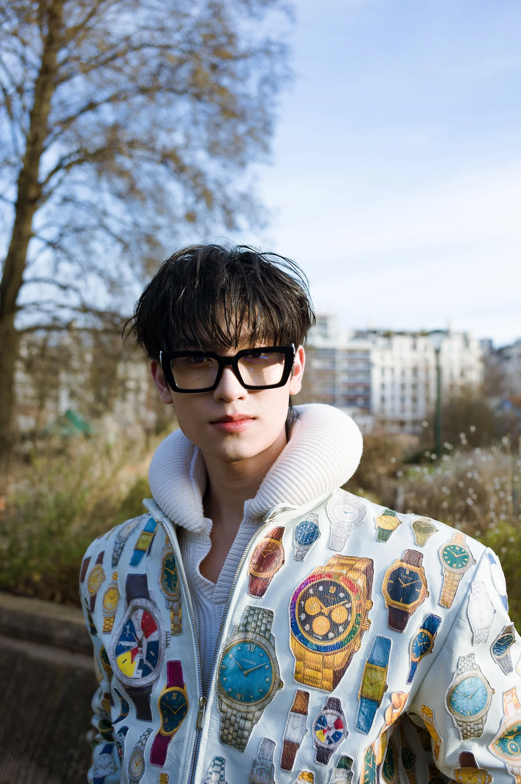 At Paris Fashion Week, A young person with short dark hair and glasses outdoors during daytime, wearing a white jacket with colorful watched designs, with trees and buildings in the background.