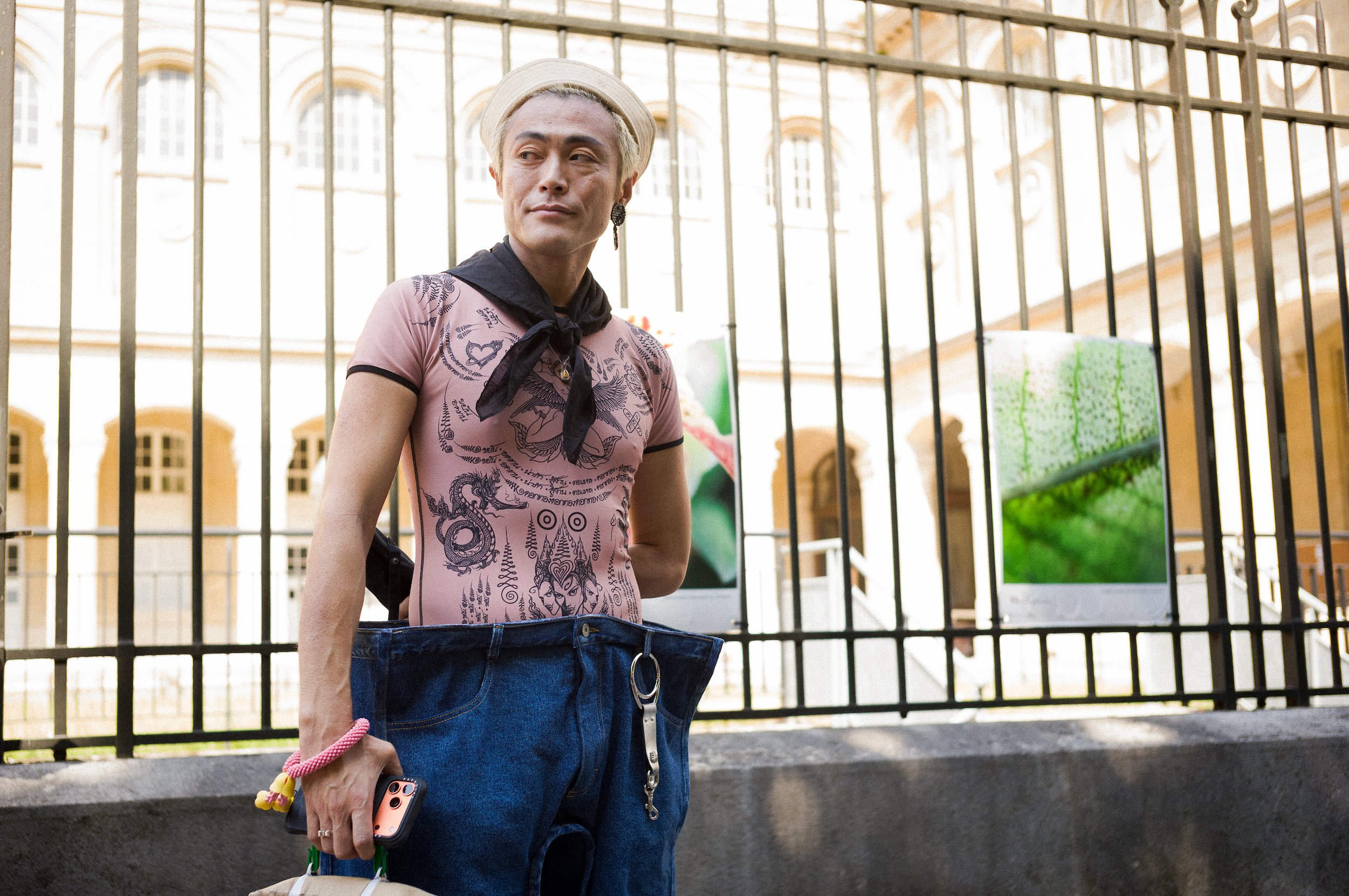 A person with platinum blonde hair wearing a pastel pink graphic T-shirt, a beige beret, and a black scarf tied around the neck stands in front of a metal fence. They are holding a phone and a shopping bag, with a building with large windows and deco