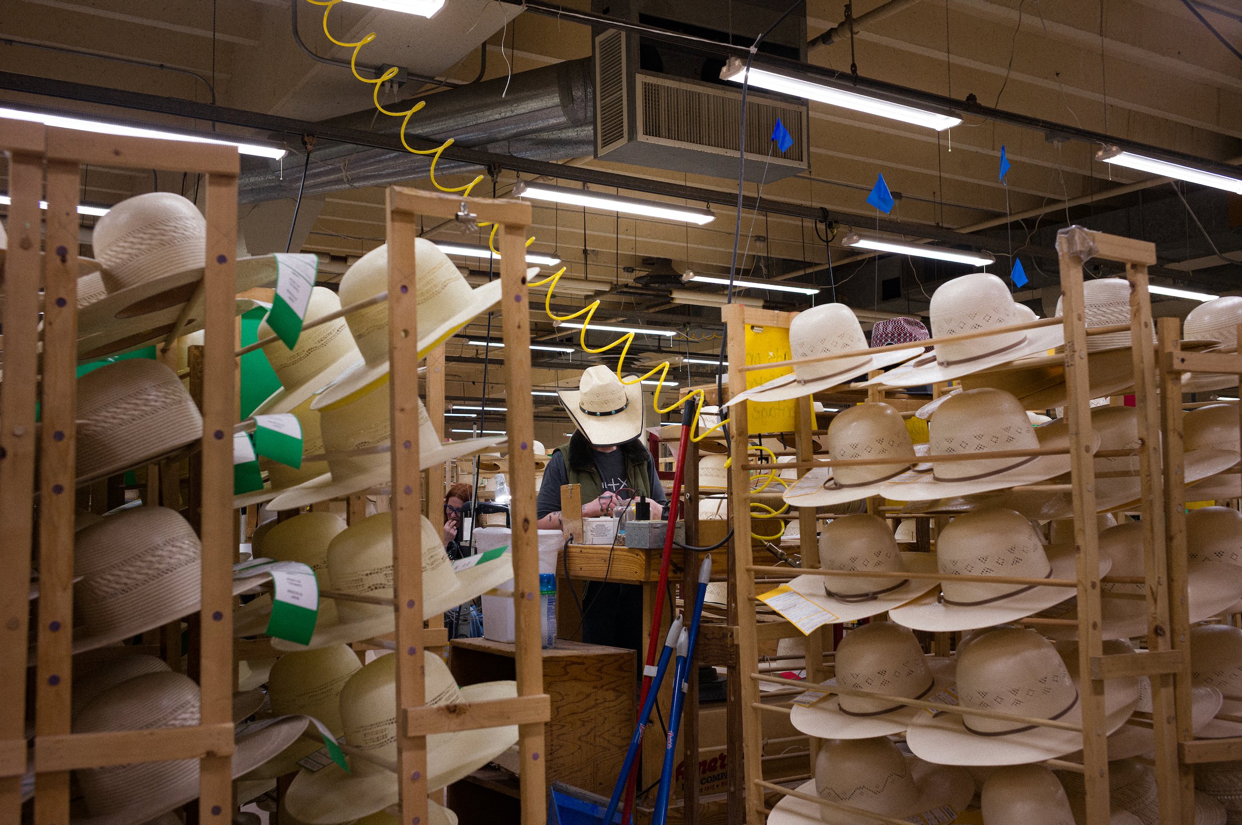 Rows of straw hats with wide brims displayed on wooden shelves in the american hat company workshop, with fluorescent lighting and industrial ceiling fixtures overhead.