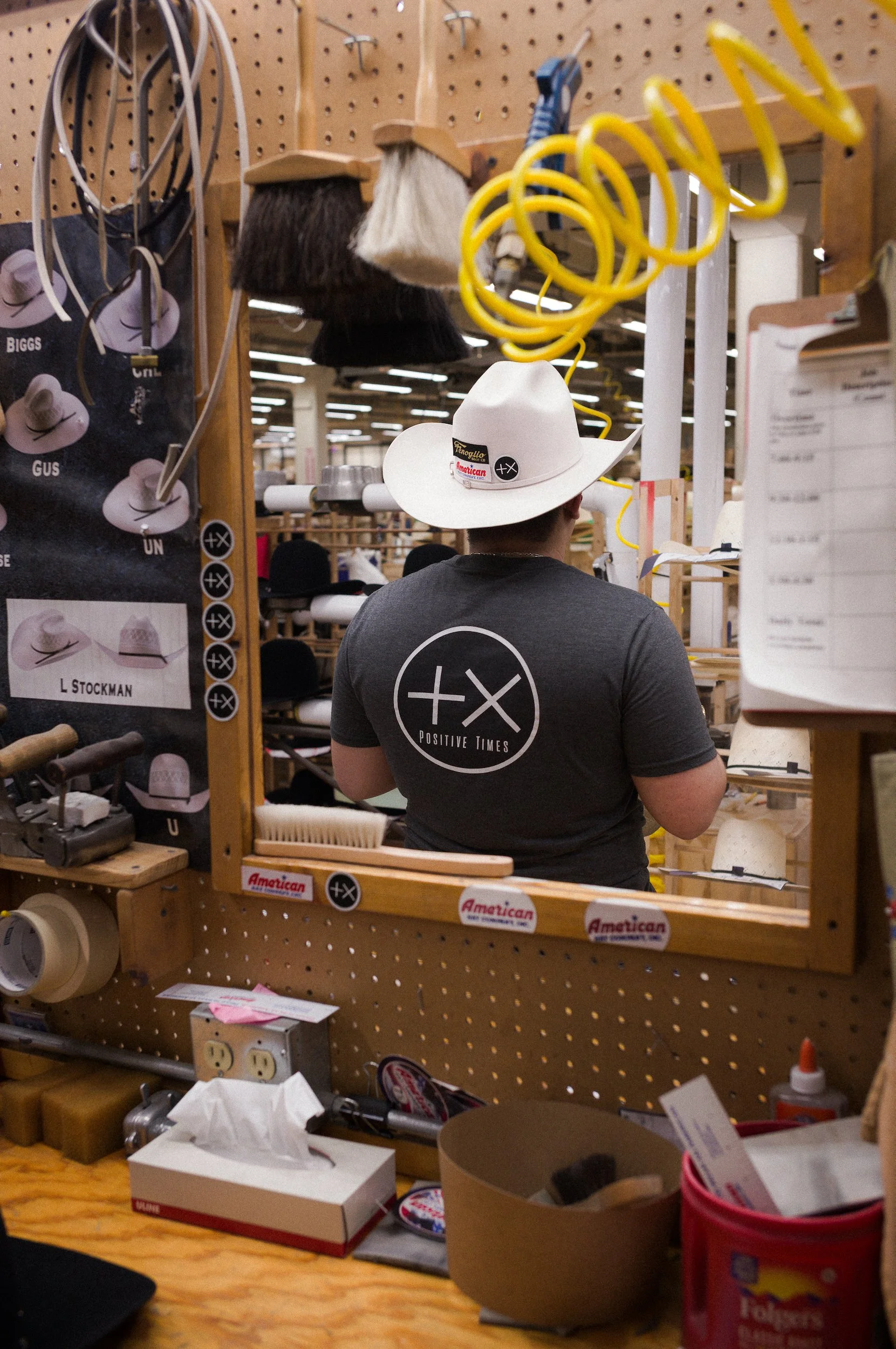 Back of a person wearing a white cowboy hat with postive times and american hat company stickers, viewed from behind, at a cluttered workspace with tools, a pegboard wall, and miscellaneous items.