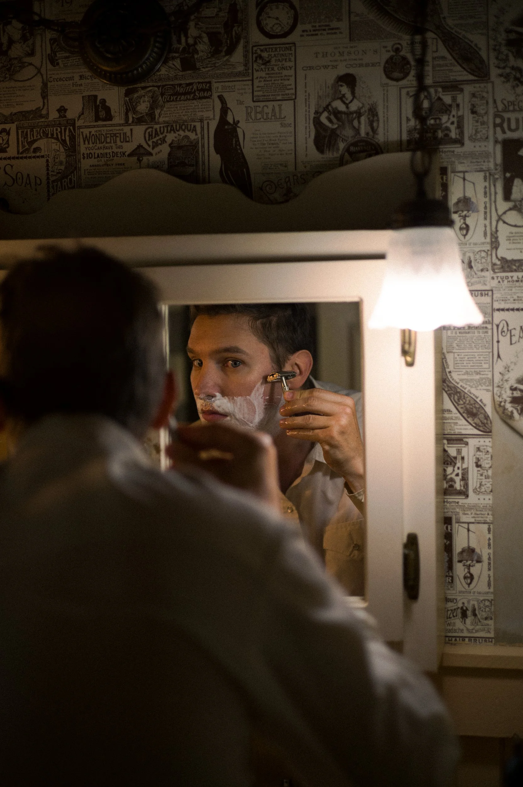 A young man shaving in front of a mirror in a vintage-style bathroom with newspaper-patterned wallpaper.