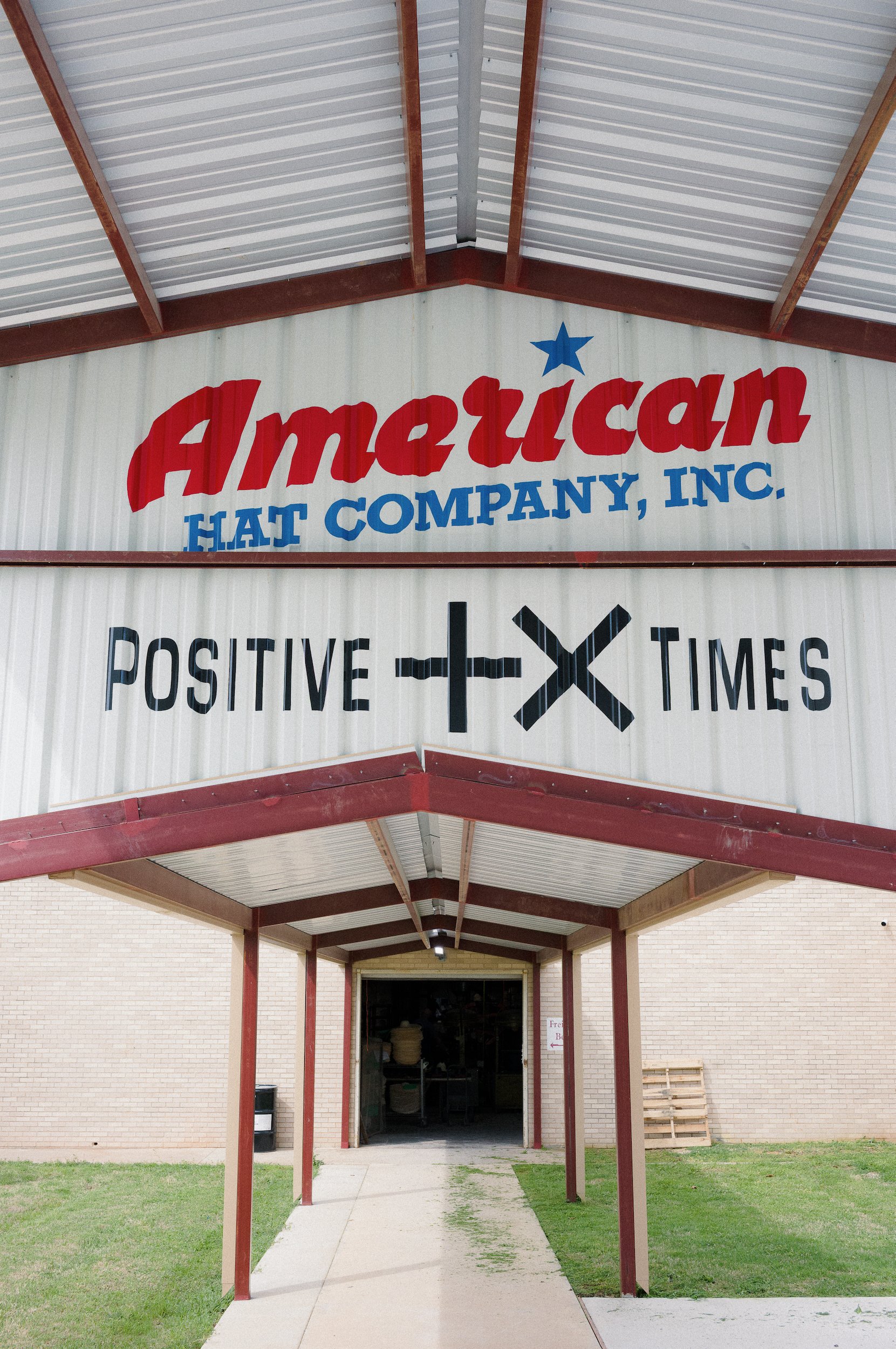 The entrance of American Heat Company, Inc. building with a sign displaying the company name and slogan, located under a metal roof supported by red poles, with a concrete walkway leading inside.