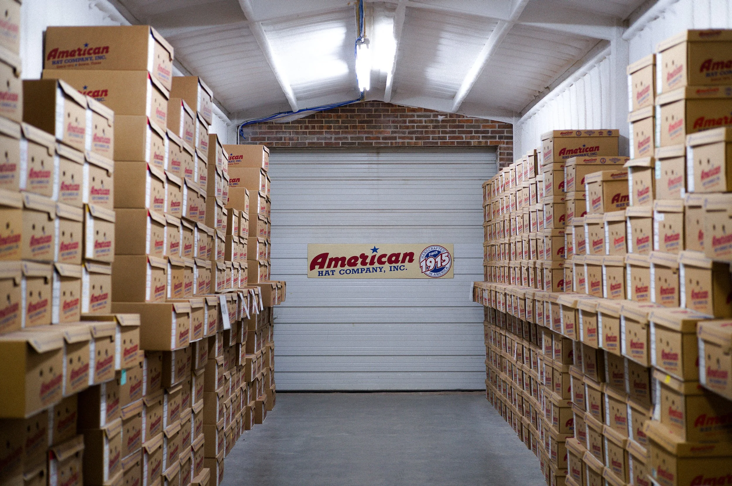A storage room with stacks of boxes labeled American Hat Company, Inc. and a sign on the back wall that says American Hat Company, Inc. since 1915.