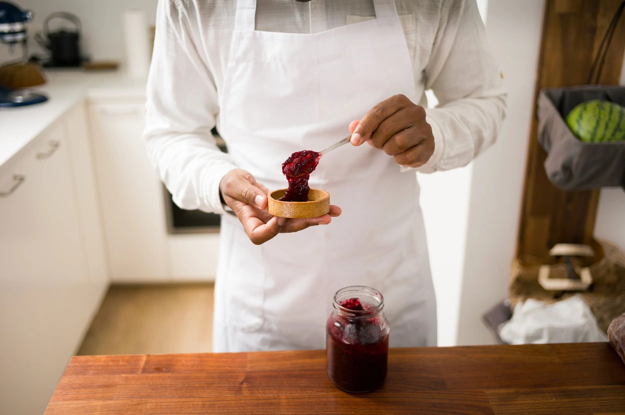 chef using a spoon to transfer homemade berry jam from a jar into a small bowl in a kitchen.